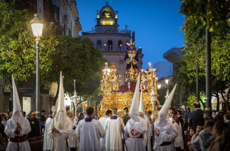 Procesión con figuras encapuchadas y carroza ornamentada al anochecer, Sevilla, España.