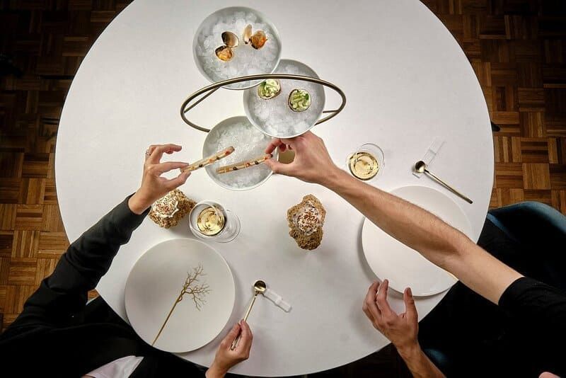 Vista aérea: personas tomando comida en una mesa blanca preparada con platos, pan e iluminación moderna.