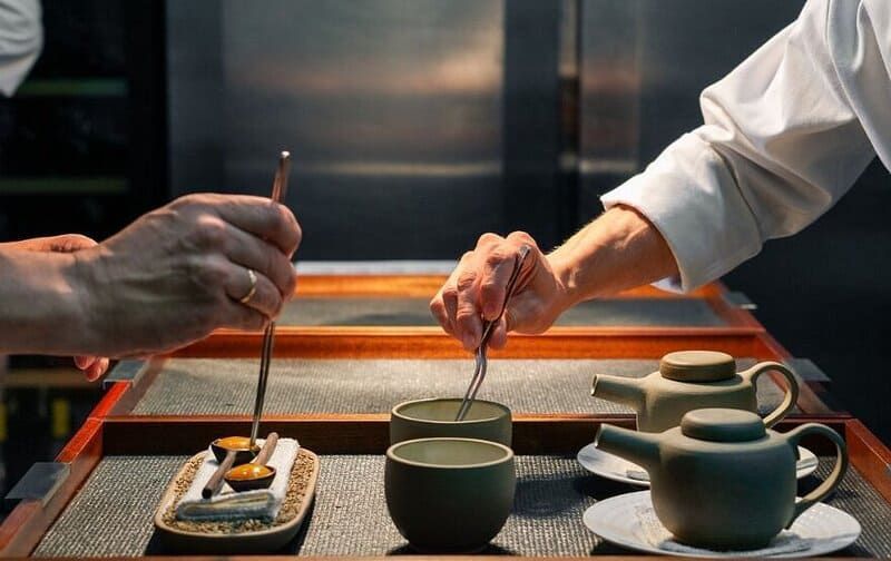 Chefs preparando comida y té en una cocina. Teteras y tazas de color verde oscuro, y una bandeja con platos pequeños.