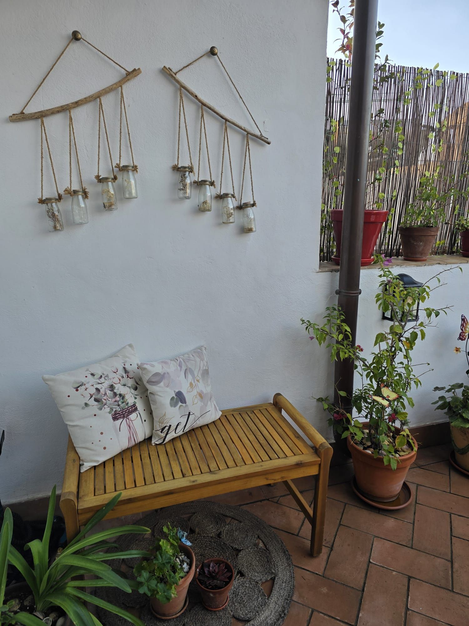 Acogedora zona de estar al aire libre con banco de madera, almohadas decorativas, frascos colgantes y plantas en macetas.