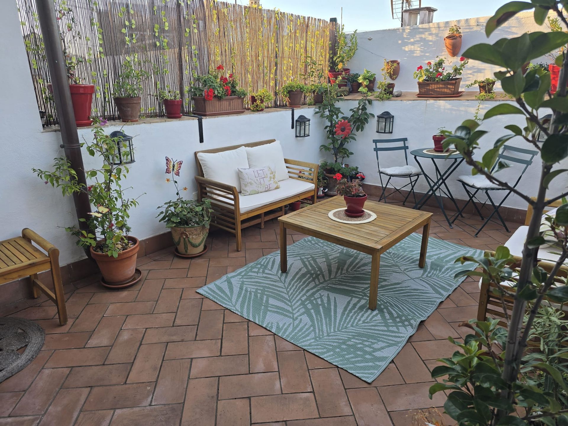 Patio exterior con muebles de madera, plantas y alfombra sobre suelo de ladrillo.