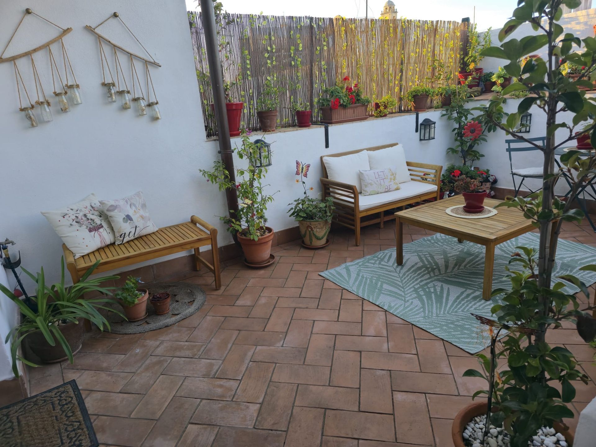 Terraza con suelo de ladrillo, muebles de madera y macetas. Paredes blancas con jardineras colgantes decorativas.
