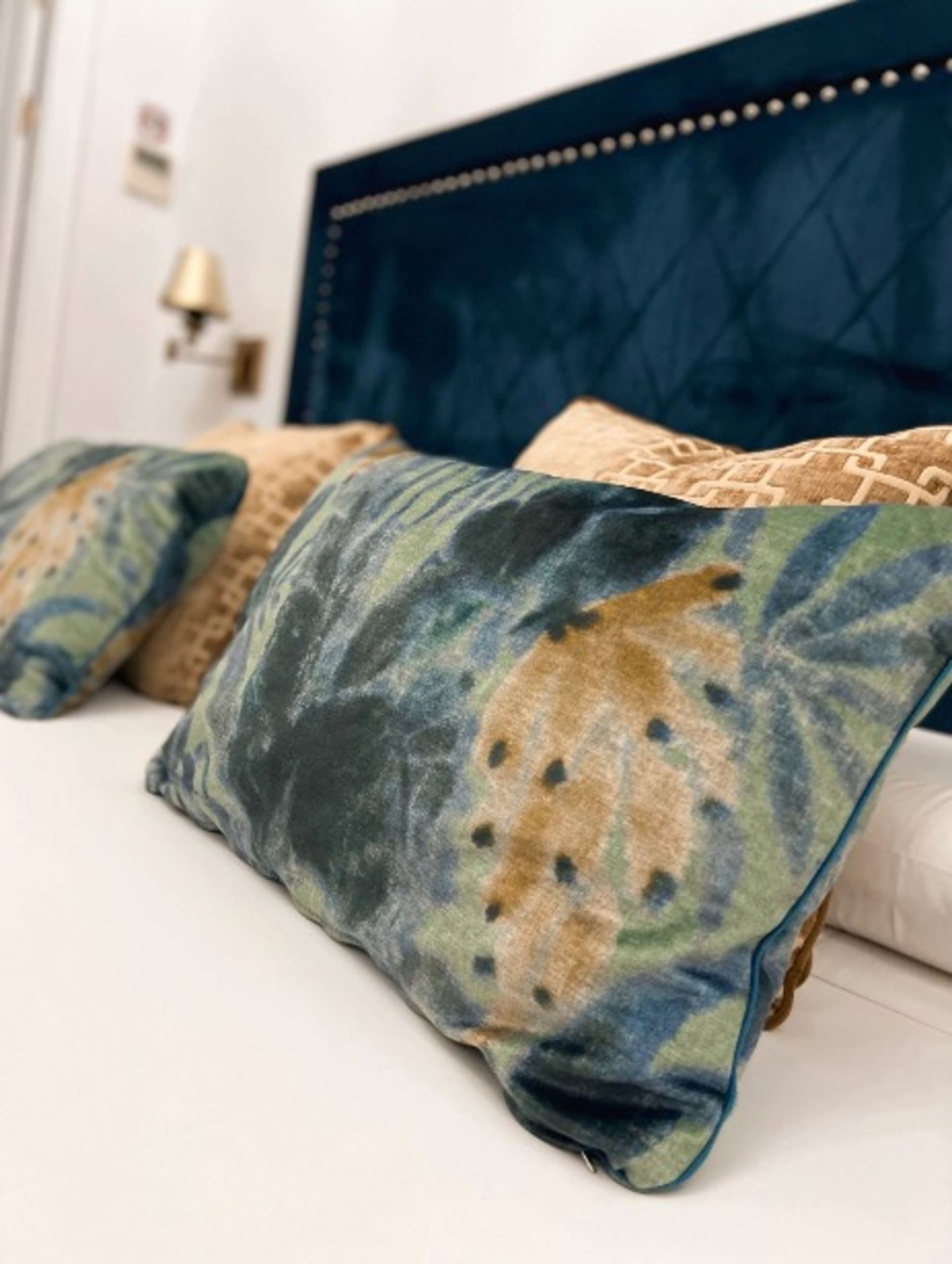 Cama con cabecero de terciopelo azul, sábanas blancas y almohadas estampadas.