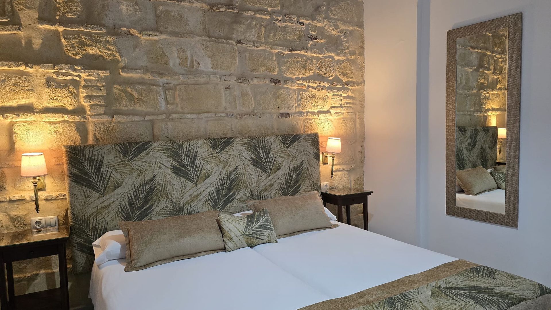 Dormitorio con pared de piedra vista, cama con cabecero y almohadas estampadas, mesas de luz y espejo.