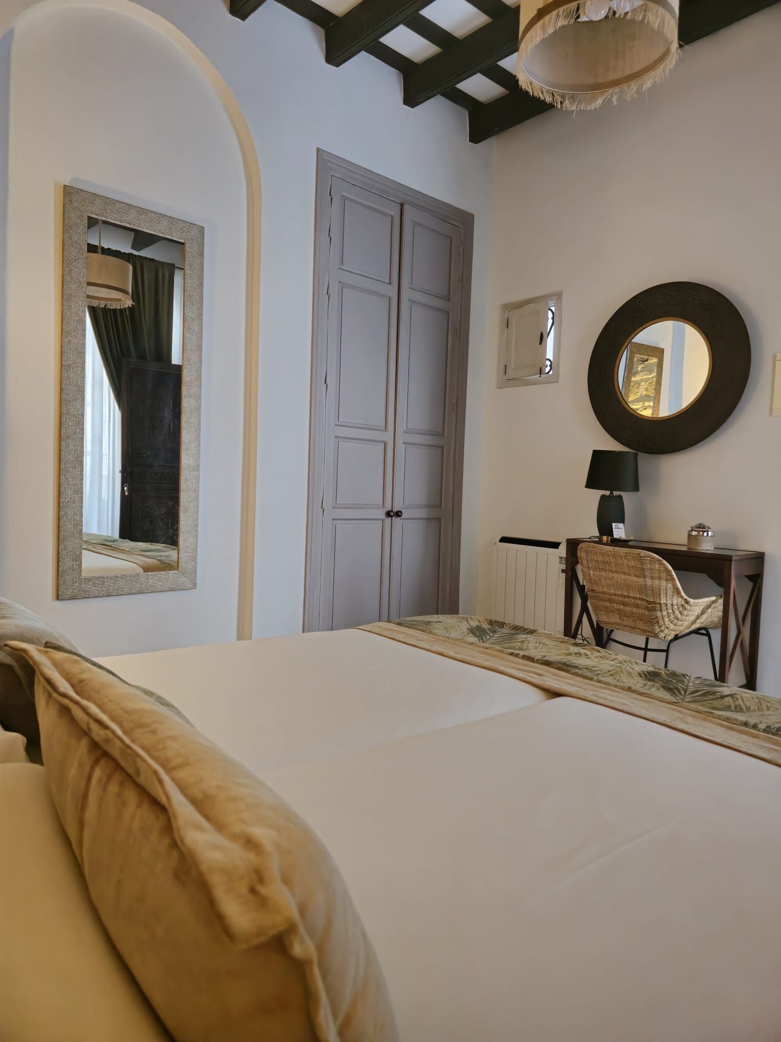 Dormitorio con paredes blancas, cama grande, puertas grises, espejo redondo, escritorio y puerta arqueada.