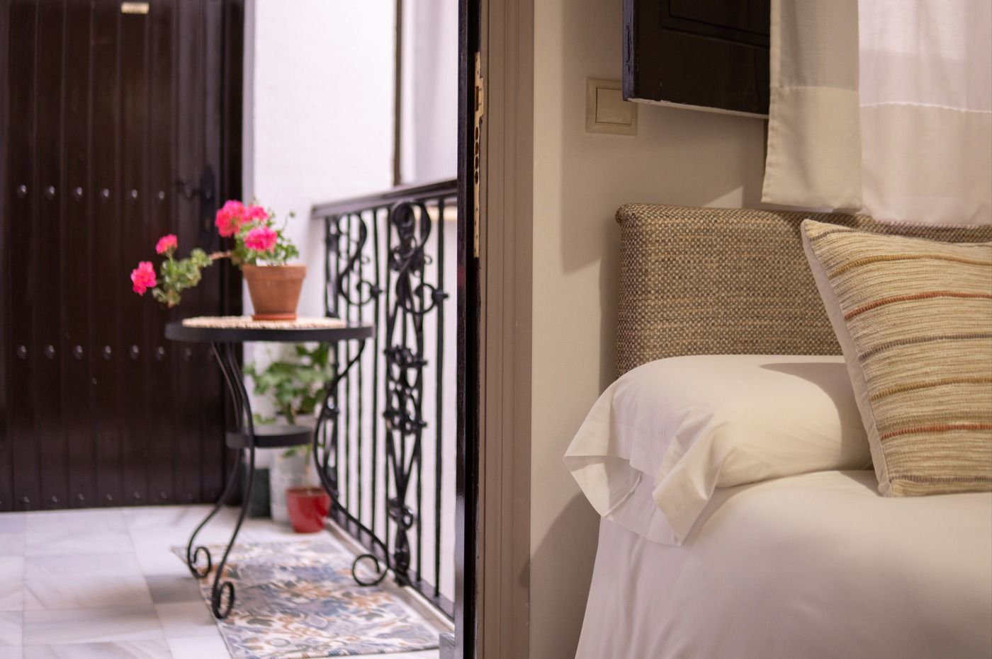 Interior de dormitorio con balcón. A la derecha hay una cama con almohadas. En el balcón hay una mesita con una maceta.