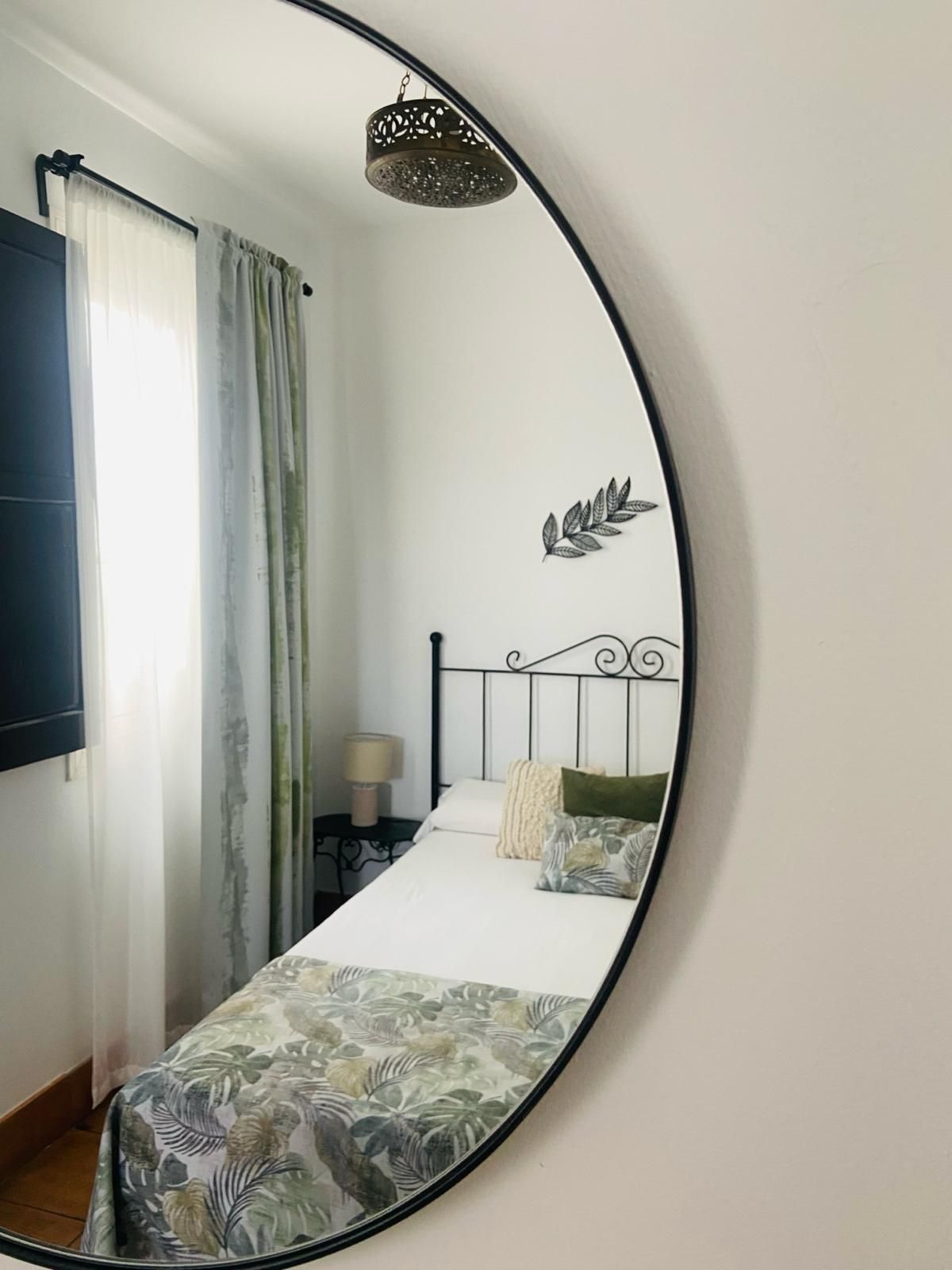 Dormitorio reflejado en un gran espejo redondo; cama, ventana con cortinas y decoración de pared.