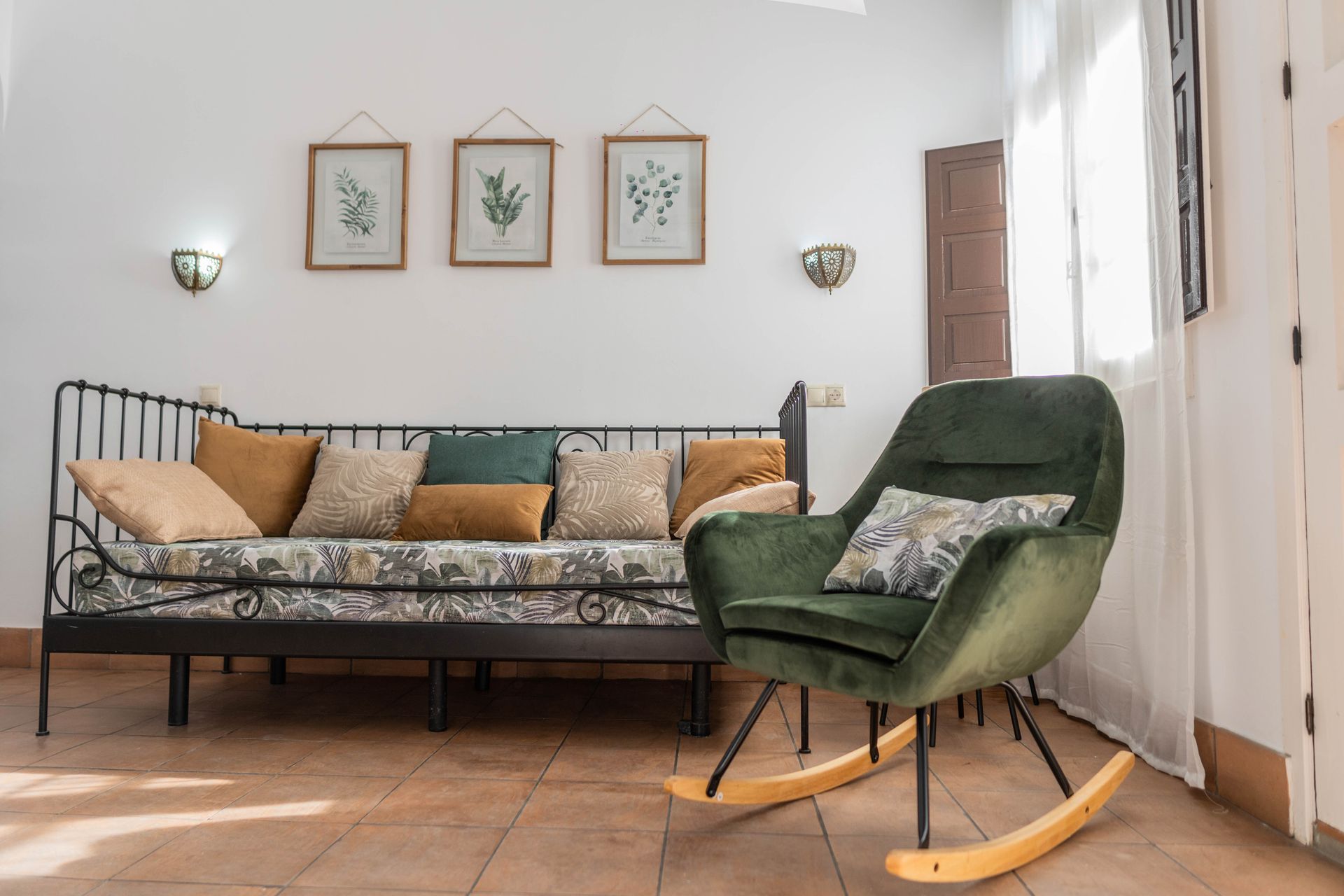 Sala de estar con mecedora verde, diván de metal negro y tres láminas botánicas enmarcadas.