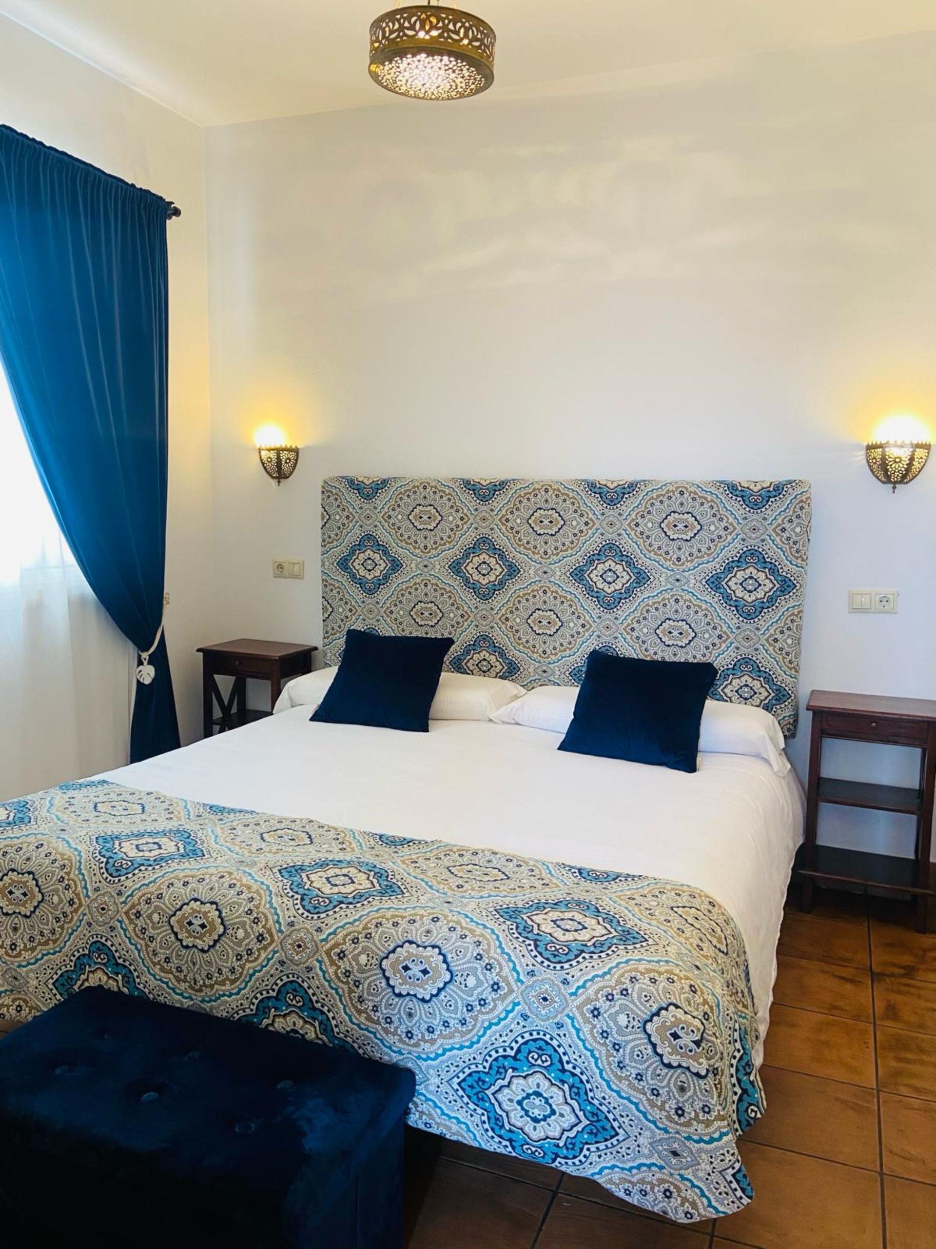 Dormitorio con cabecero, ropa de cama y cortinas con estampado azul y blanco. Dos mesitas de noche, apliques y un banco.