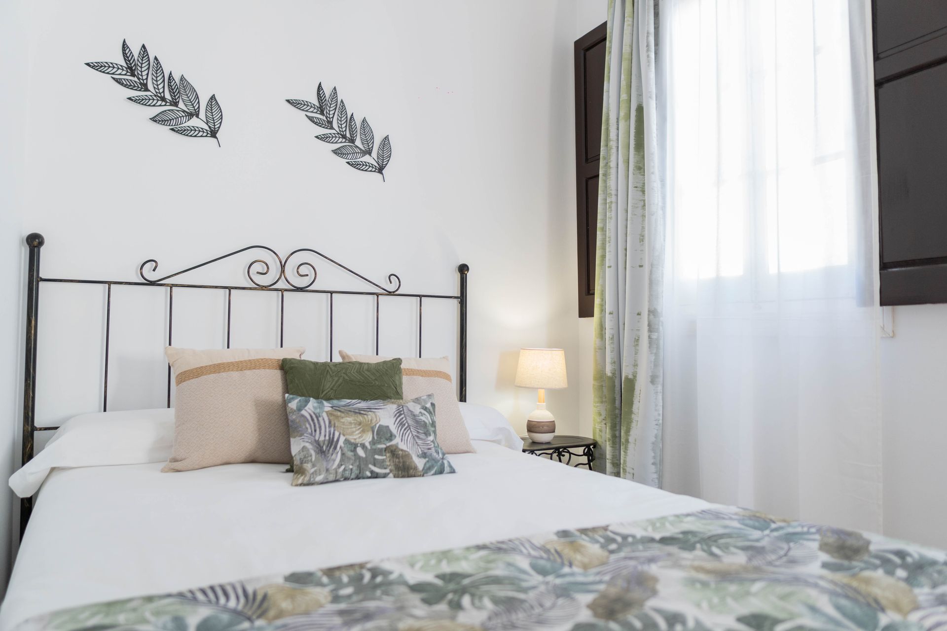 Dormitorio con paredes blancas, marco de cama de hierro, almohadas florales y ventana con cortinas transparentes.