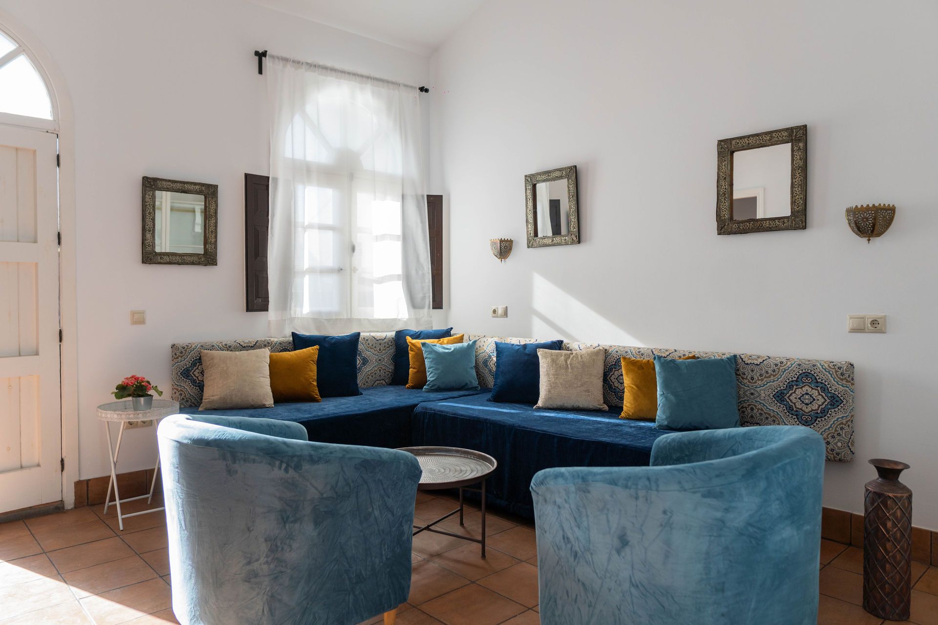 Sala de estar con sofá seccional azul, sillones y cojines decorativos. Paredes blancas, ventana y mesa pequeña.