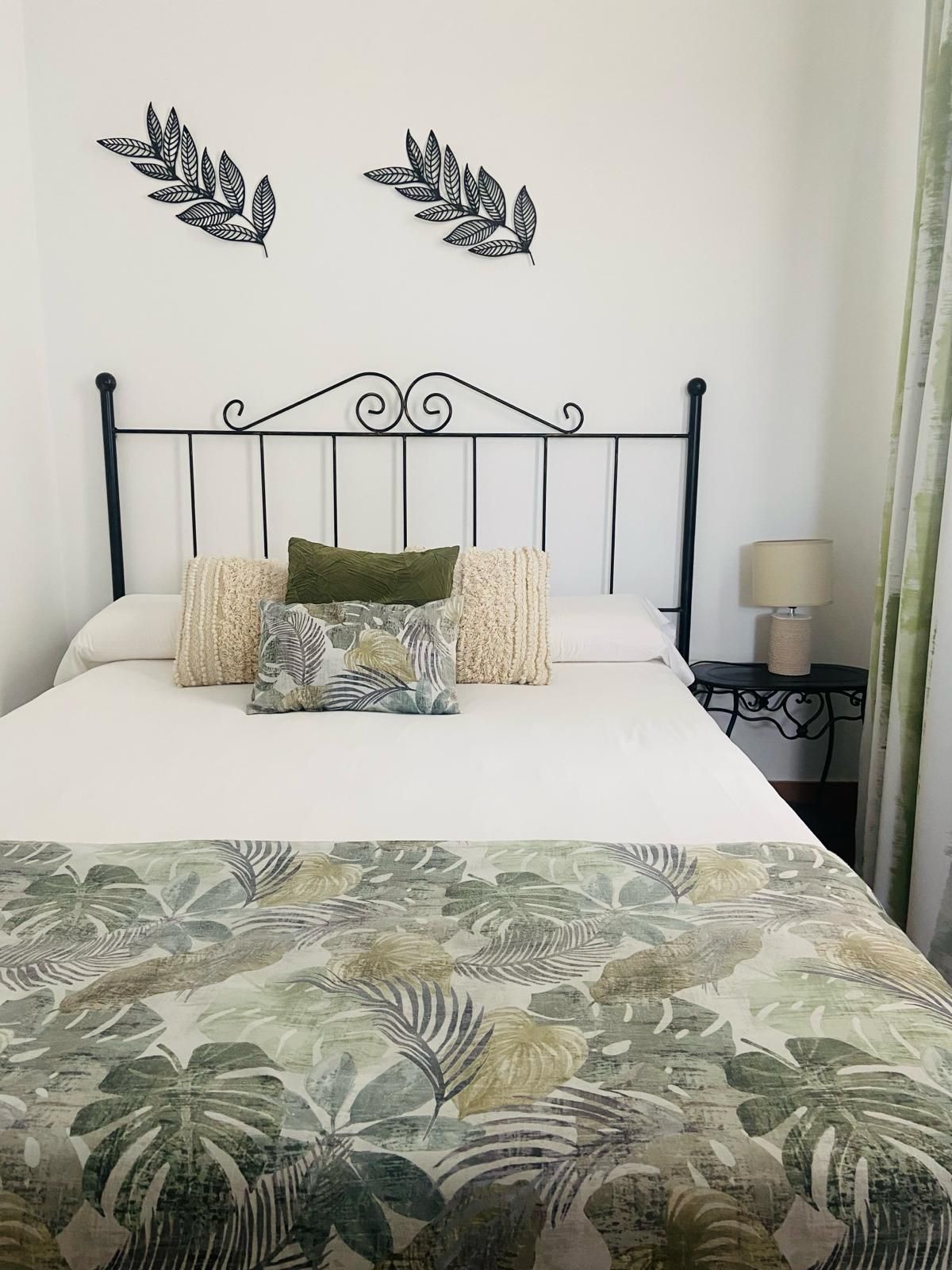 Dormitorio con cama. Cabecero de hierro forjado negro, manta floral, cojines decorativos y una lámpara pequeña.