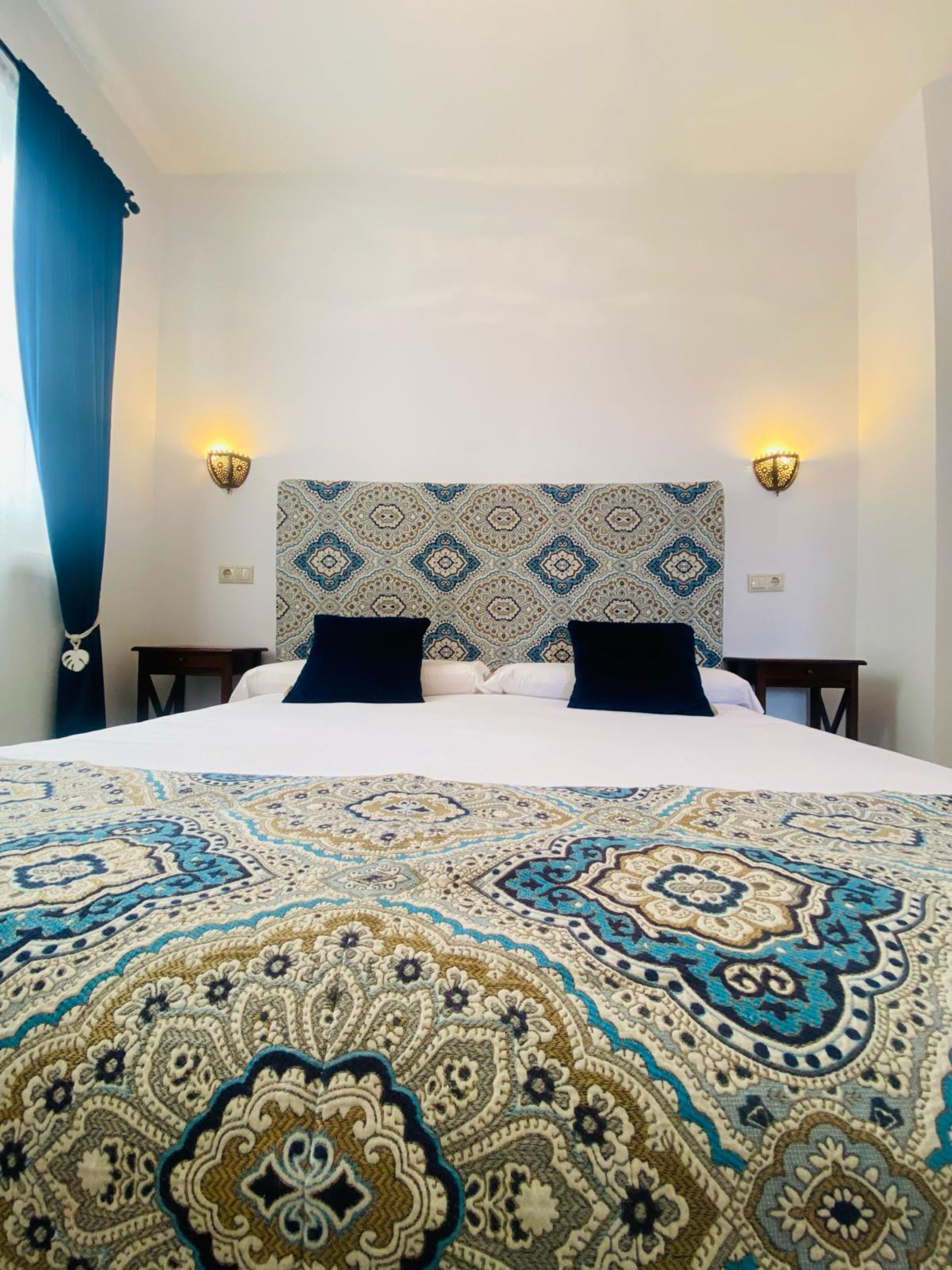Dormitorio con cama grande, cabecero y ropa de cama estampados, dos mesitas de noche, apliques de pared y cortinas azules.