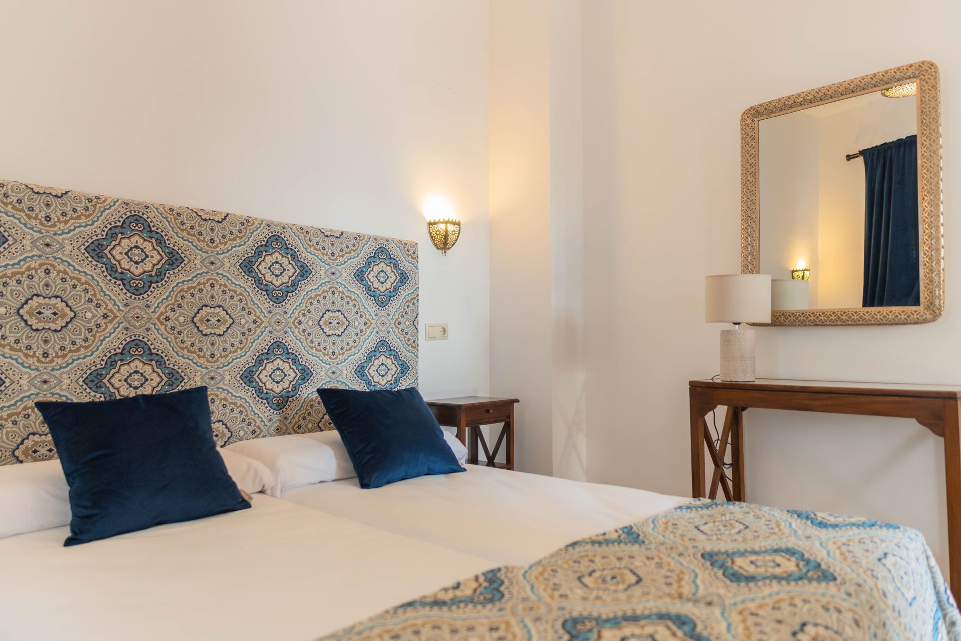 Dormitorio con dos camas, cabecero y almohadas estampadas, espejo decorativo y mesita de noche.