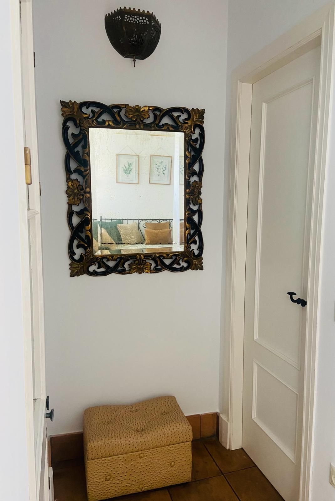 Entrada con espejo ornamentado, otomana tejida y puerta blanca.
