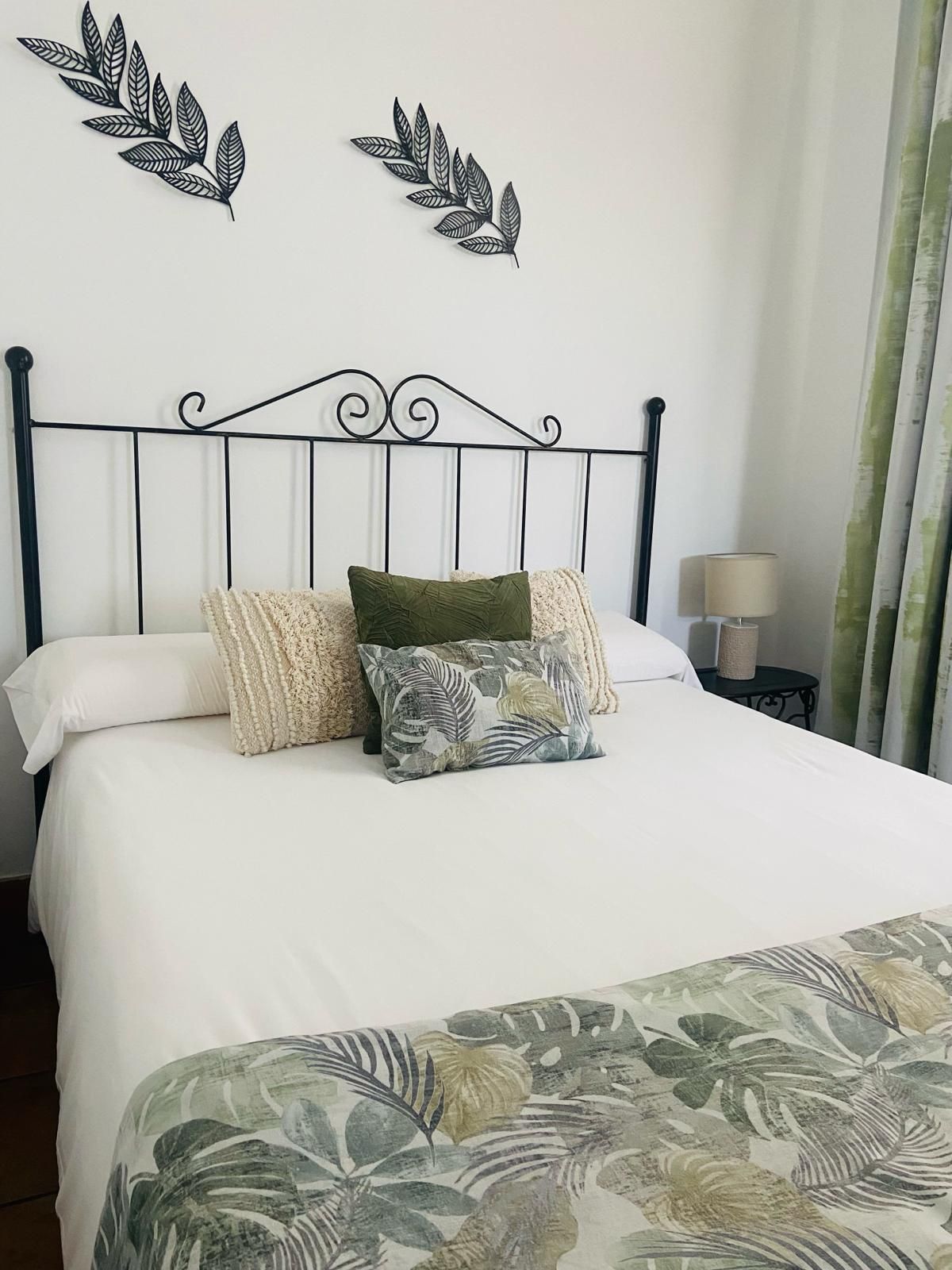 Dormitorio con ropa de cama blanca, cojines decorativos y manta con estampado floral. Cabecero negro y decoración de pared.