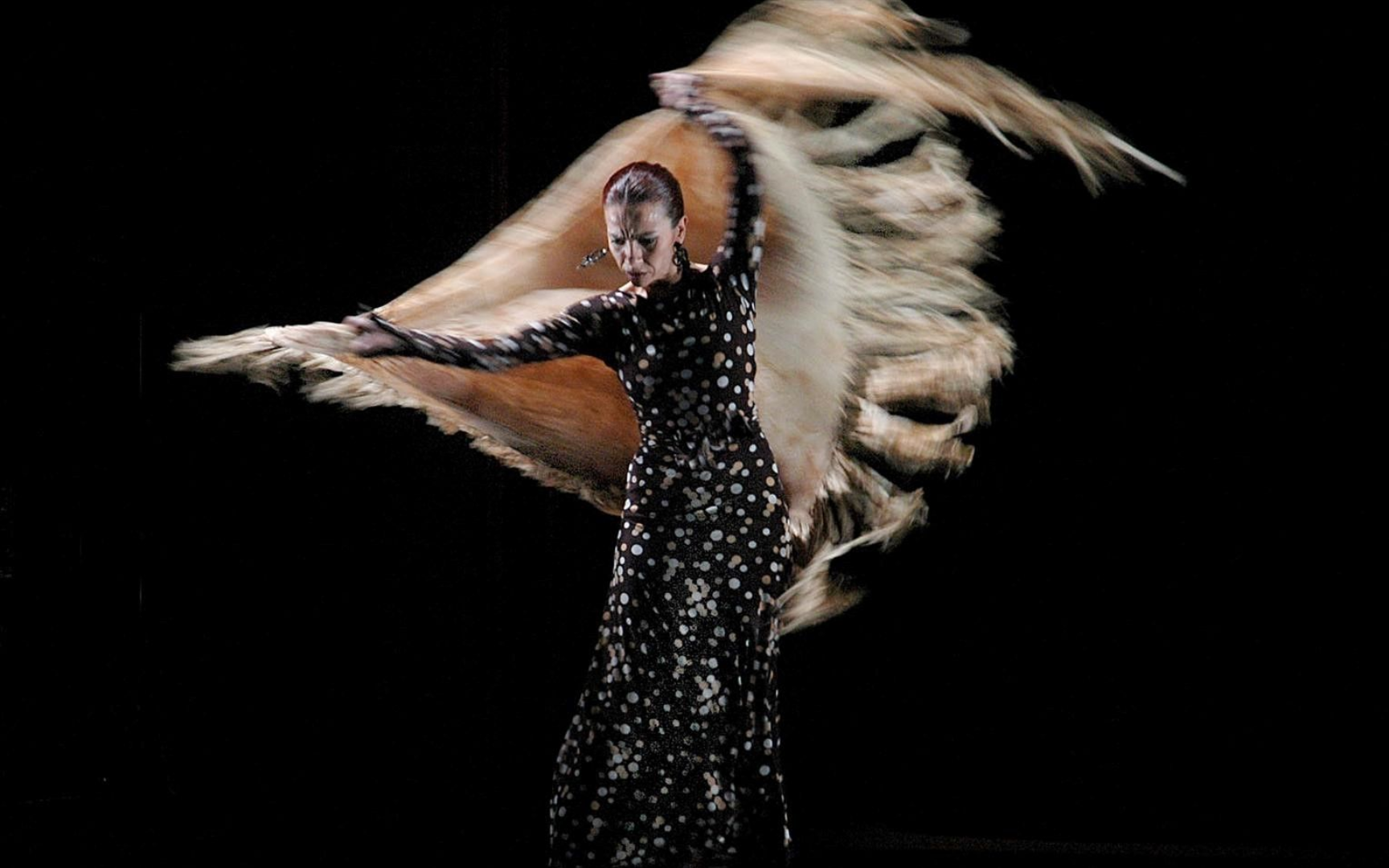 Mujer con un vestido floral haciendo girar un chal fluido con flecos sobre un fondo oscuro.