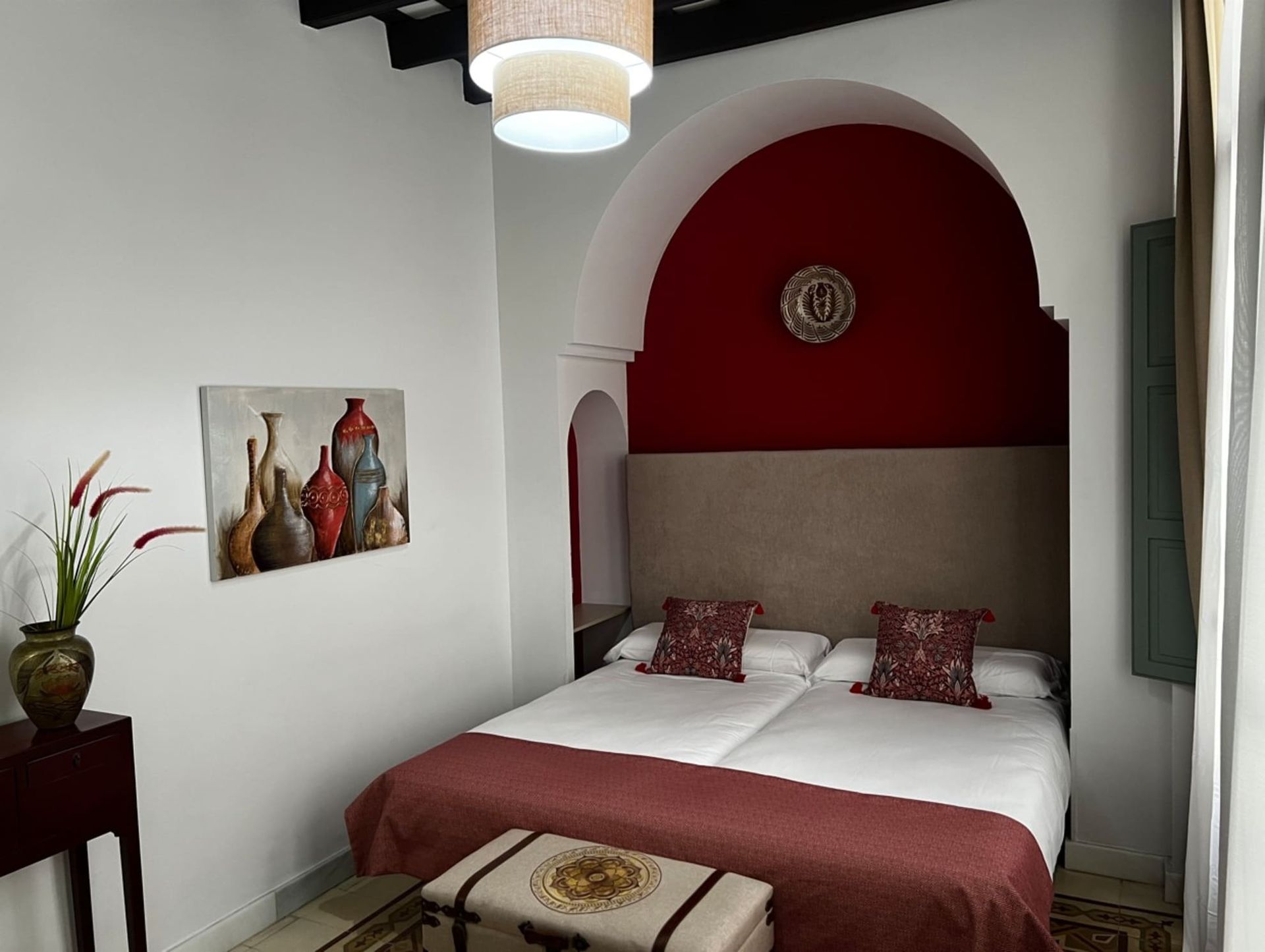 Dormitorio con alcoba arqueada, cabecera roja, dos camas individuales, almohadas decorativas, arte, mesa pequeña y baúl.