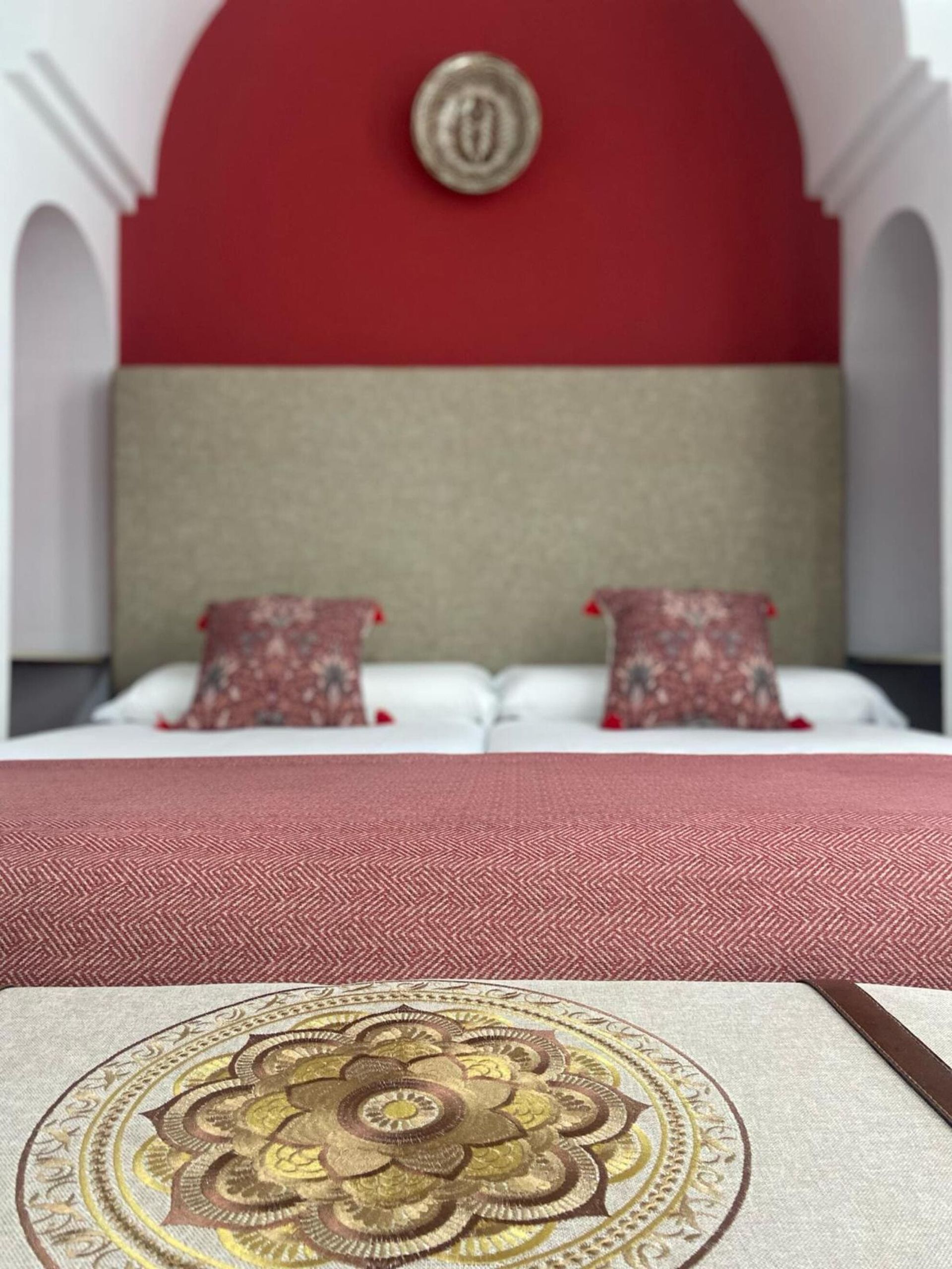 Dormitorio con detalles en rojo, arcos y una cama con almohadas decorativas y un colgante de pared de color dorado.