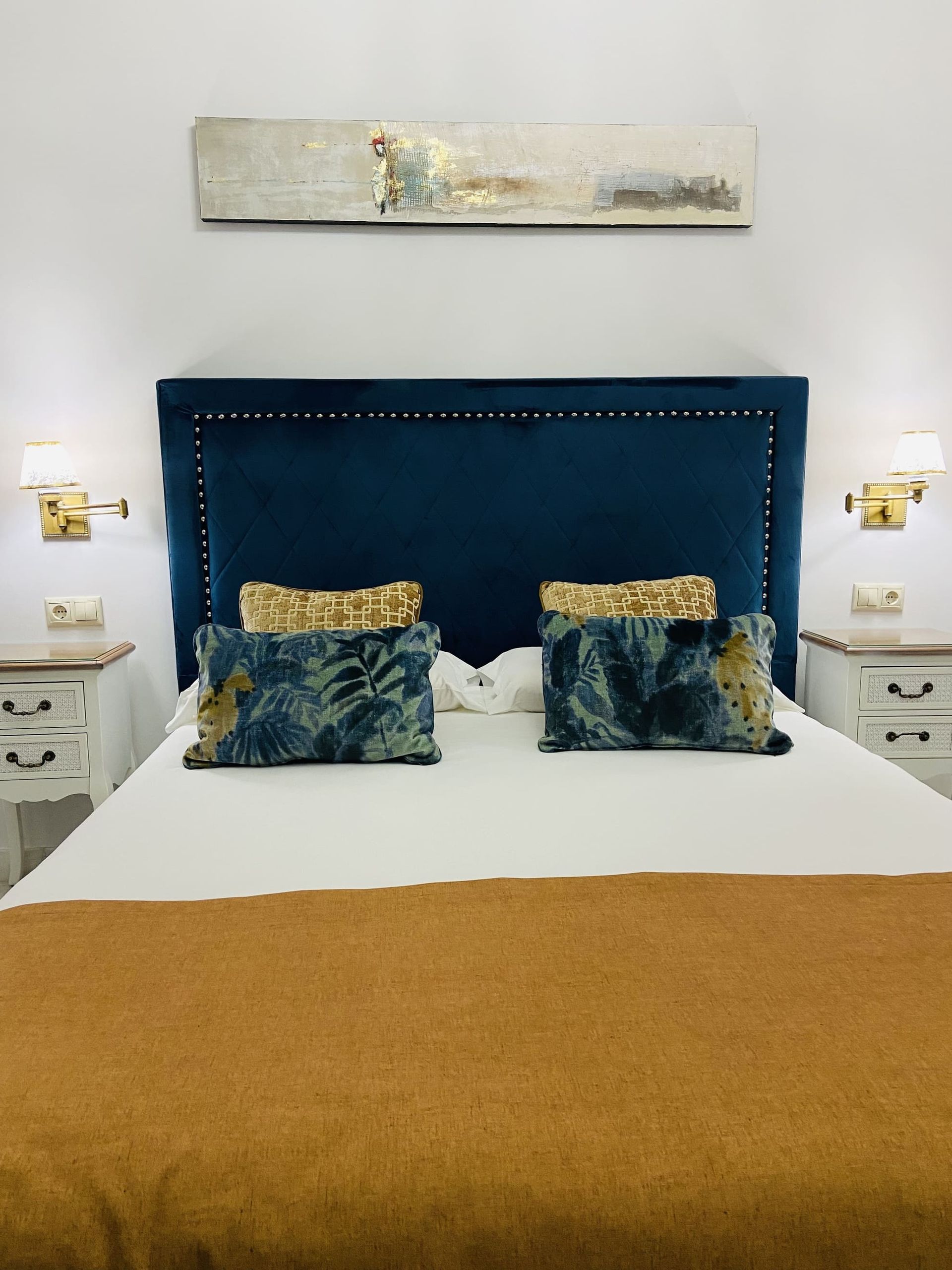 Cabecera de terciopelo azul con detalles dorados, cama con manta dorada, paredes blancas, mesitas de noche.