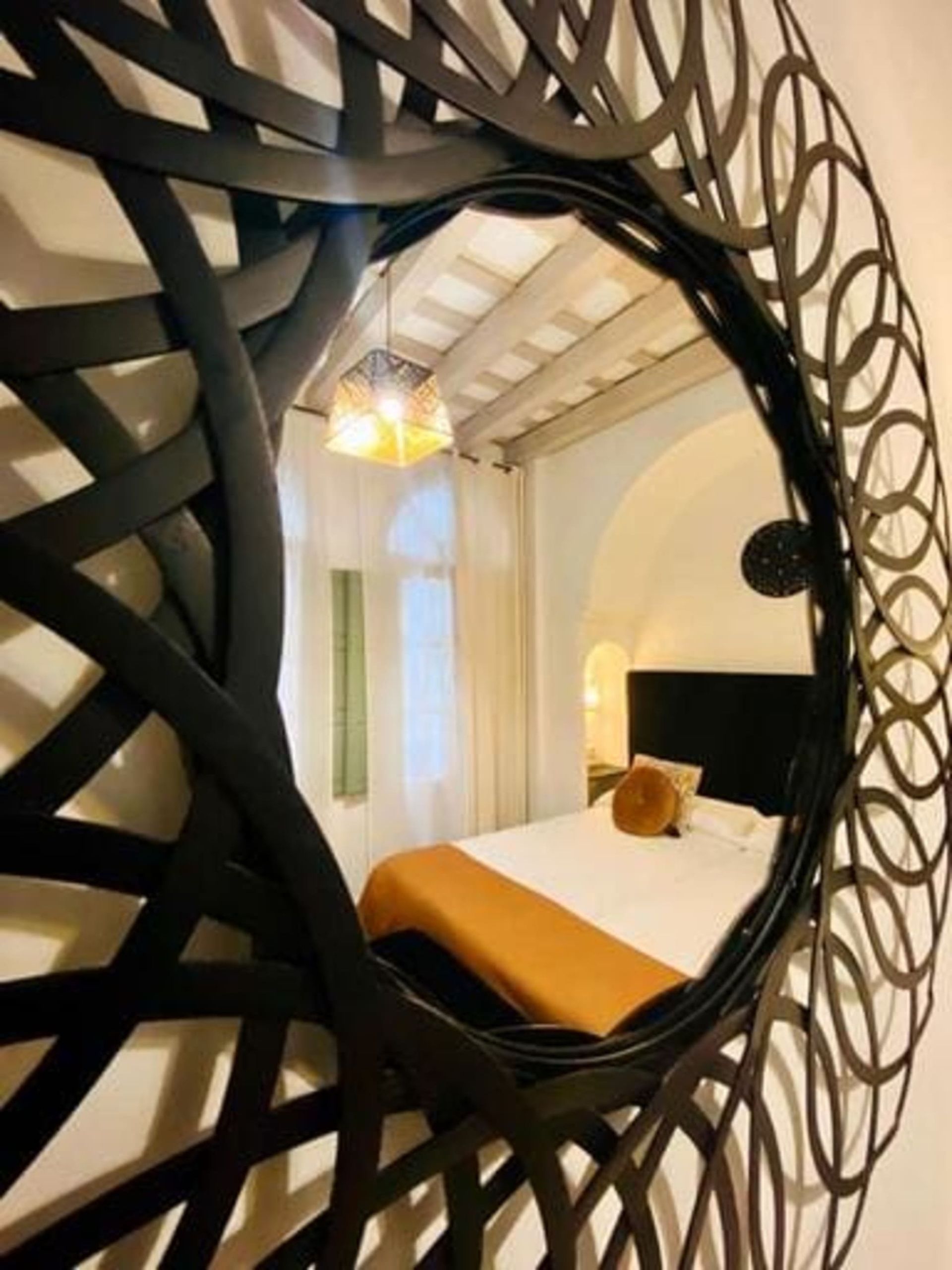 Espejo que refleja un dormitorio con cama, ventana e iluminación decorativa. El espejo tiene un marco oscuro y elaborado.