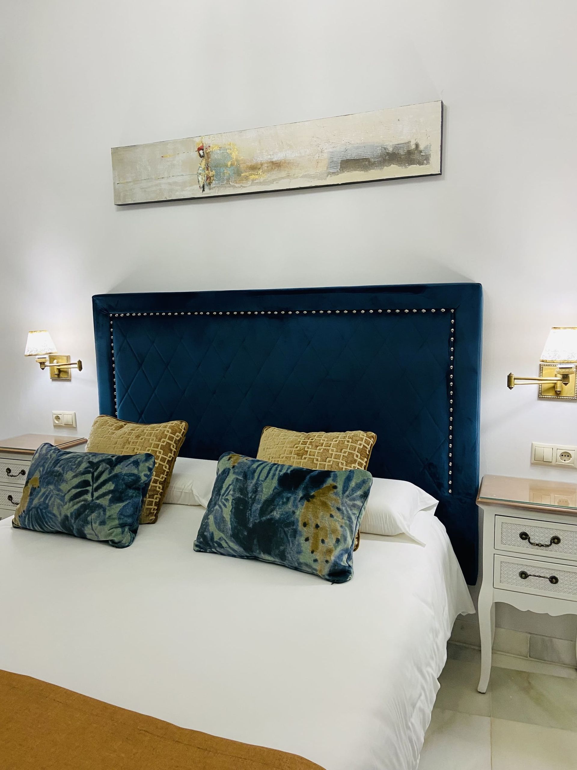 Dormitorio con cabecera de terciopelo azul, ropa de cama blanca, detalles dorados, dos mesitas de noche.