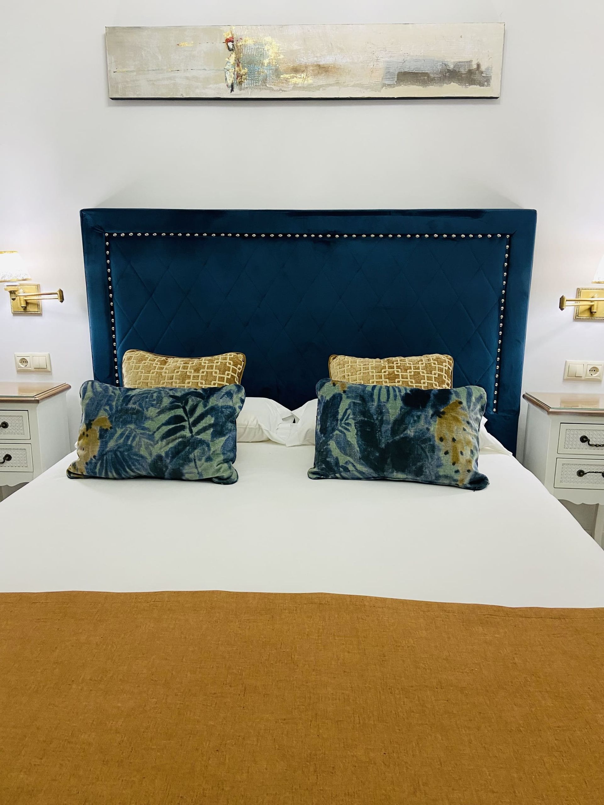 Dormitorio con cabecera de terciopelo azul, ropa de cama blanca, detalles dorados y obras de arte.