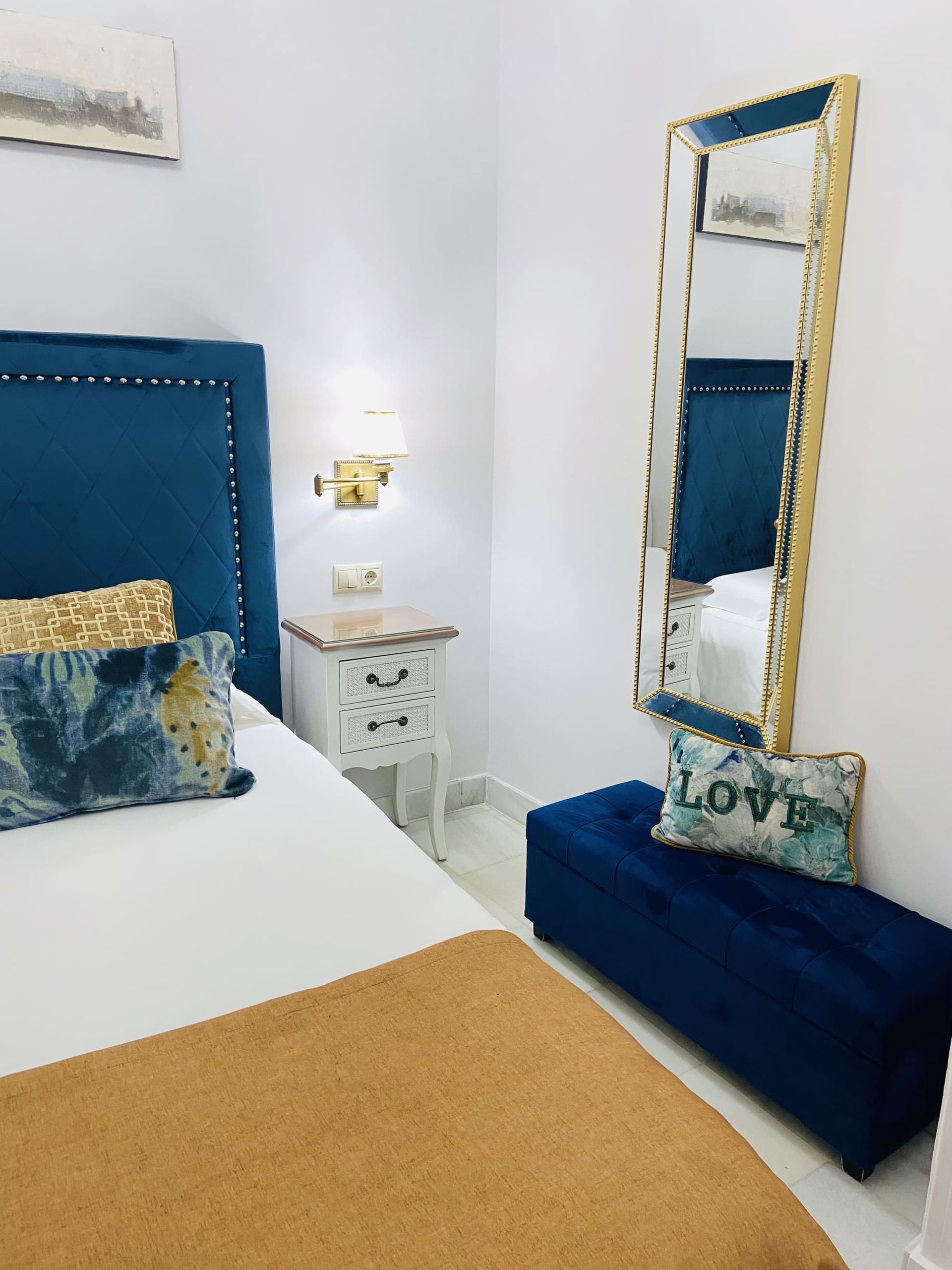 Dormitorio con cabecera de terciopelo azul, espejo con marco dorado.