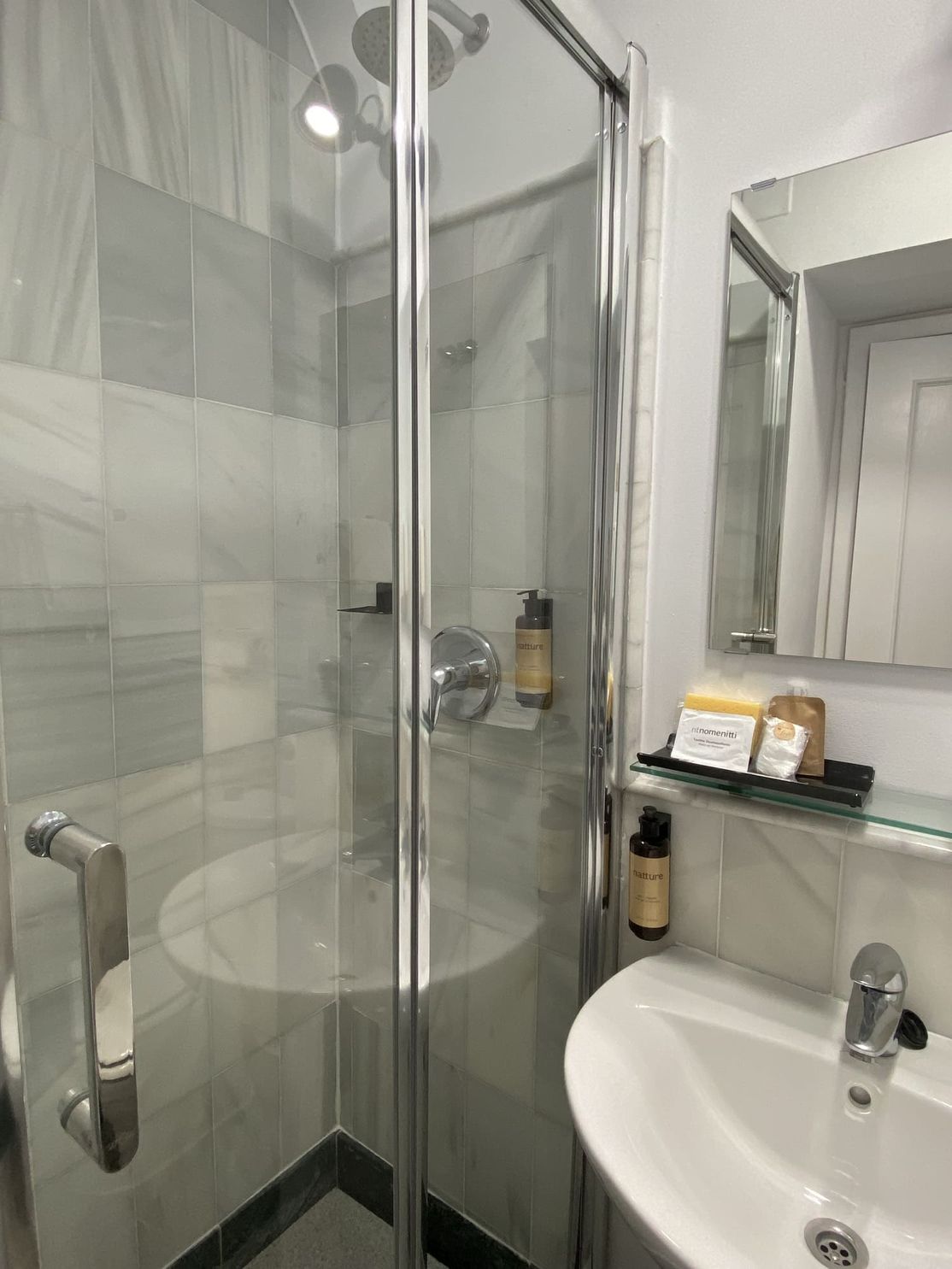 Una pequeña ducha con mampara de cristal junto a un lavabo y un espejo. Las paredes están alicatadas en gris claro.