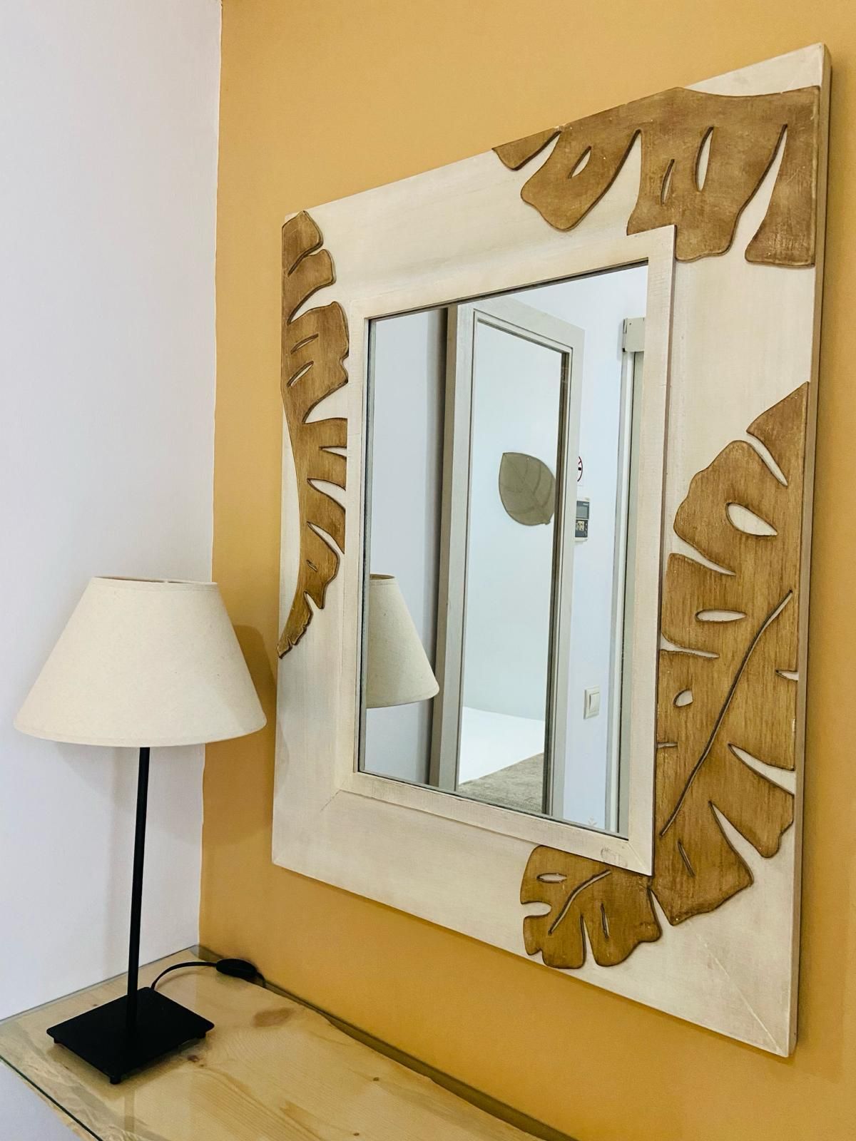 Espejo con marco con diseño de hojas sobre pared amarilla, junto a una mesa con lámpara.