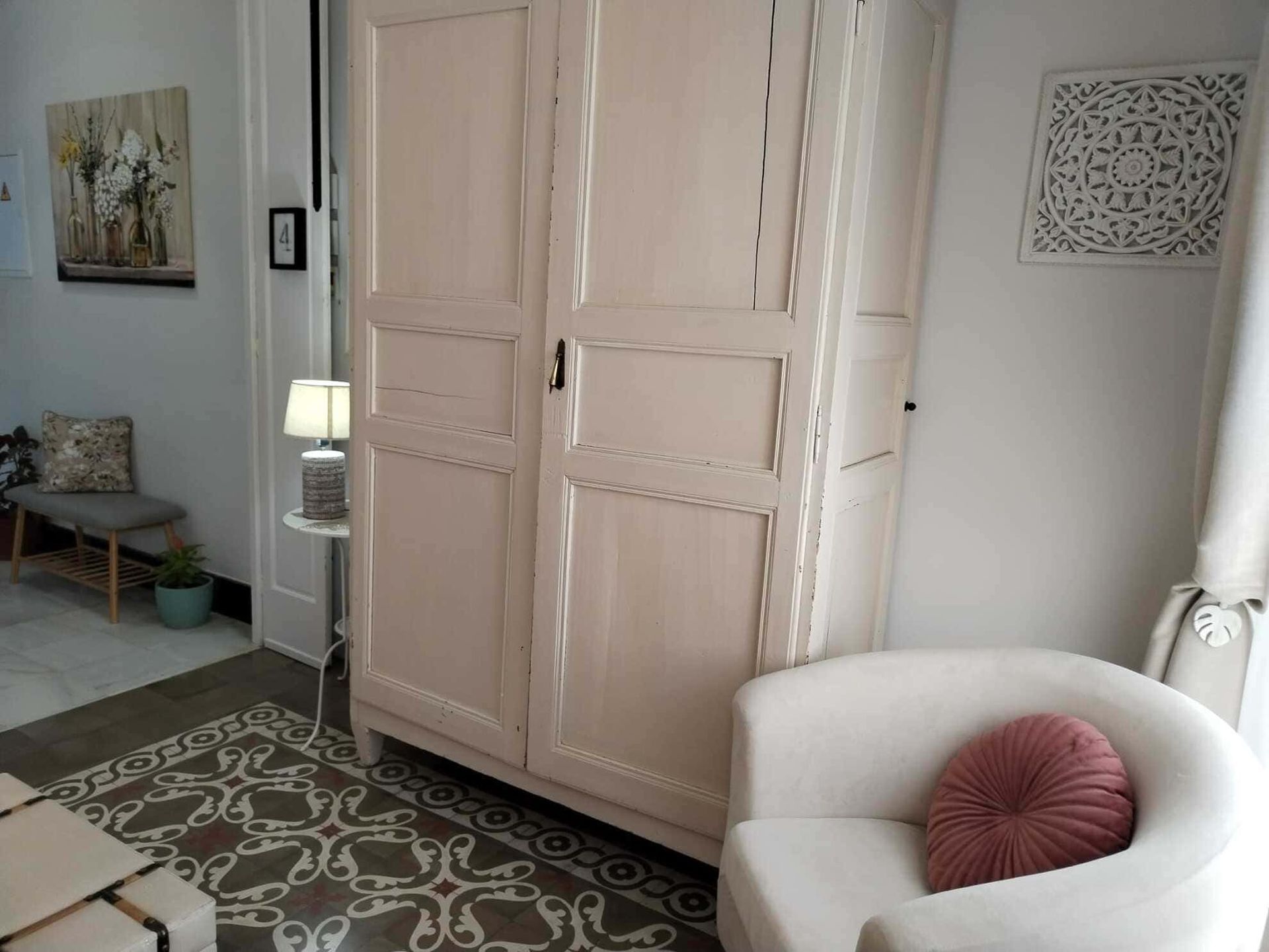 Al lado de un sillón blanco hay un armario blanco con una almohada rosa; al lado del armario hay una lámpara de mesa.