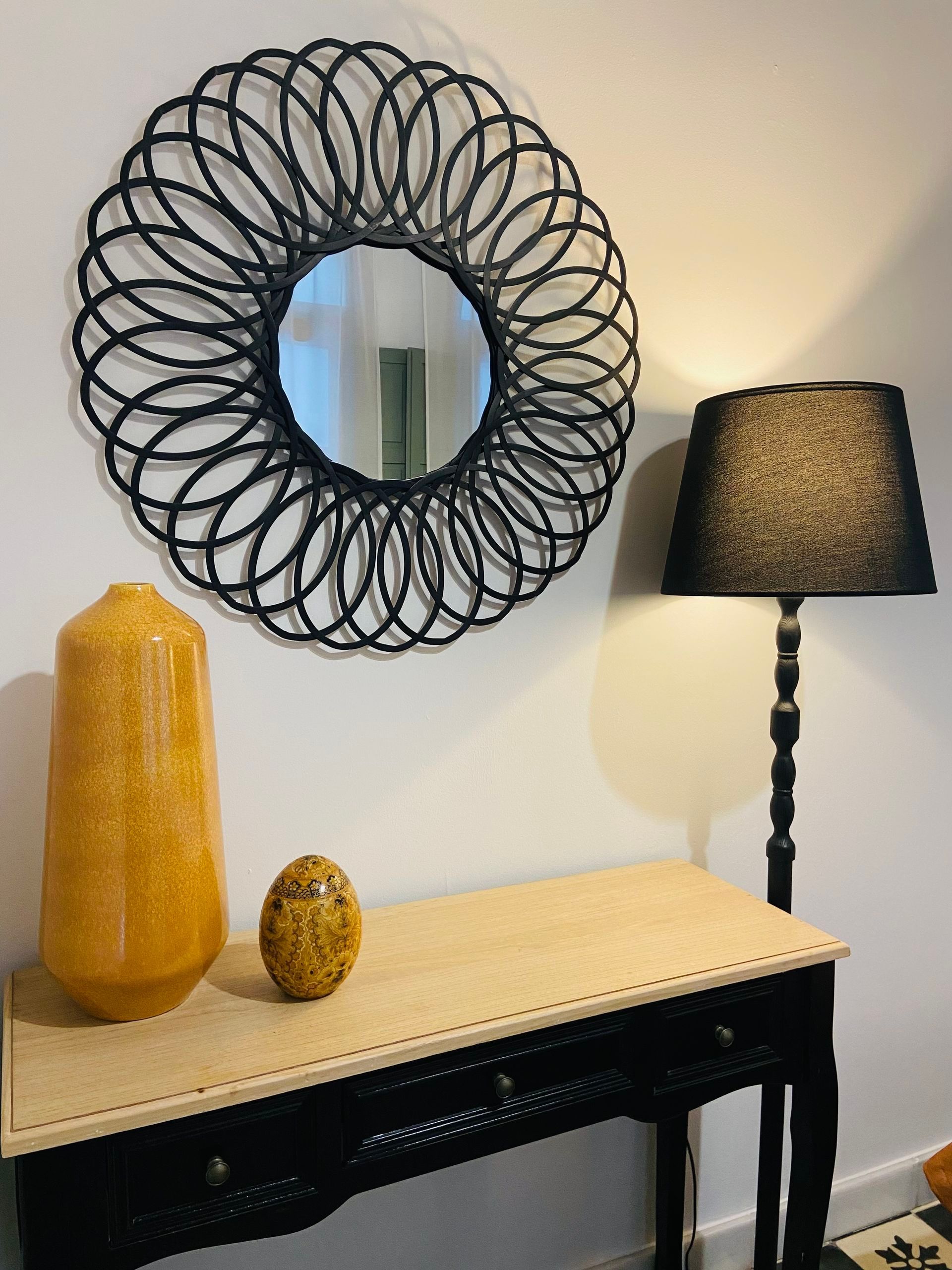 Espejo redondo con marco negro, lámpara y jarrones decorativos sobre una mesa consola contra una pared blanca.
