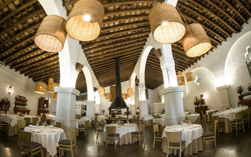 Interior de restaurante con techo abovedado, columnas blancas, mesas redondas con manteles blancos y lámparas tejidas.