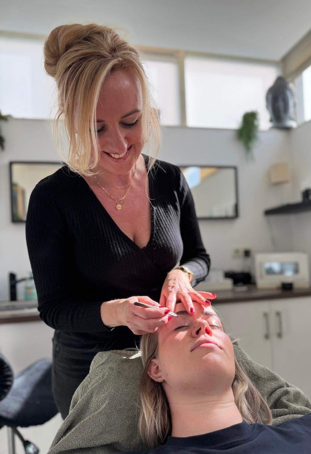 Een vrouw voert een gezichtsbehandeling uit bij een andere vrouw. Beiden bevinden zich in een salonachtige omgeving.