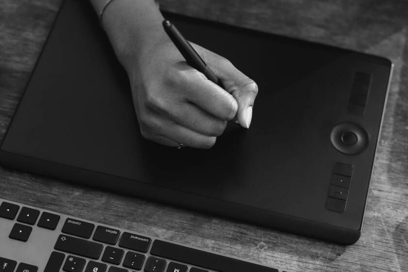 Hand using a pen on a black drawing tablet, near a keyboard, on a wooden surface.