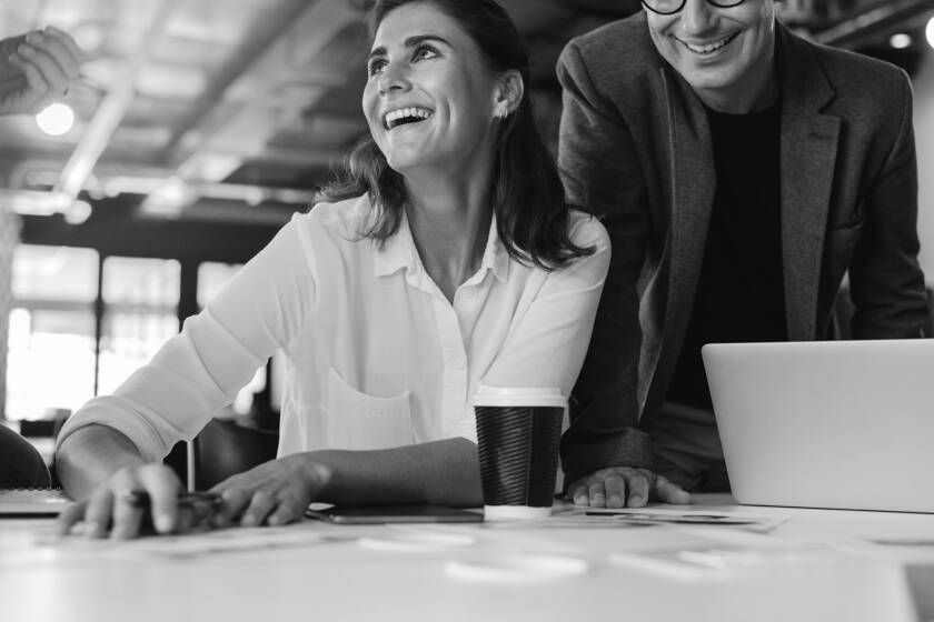 Woman smiling with a man; both look at laptop on table. Office setting, black and white.