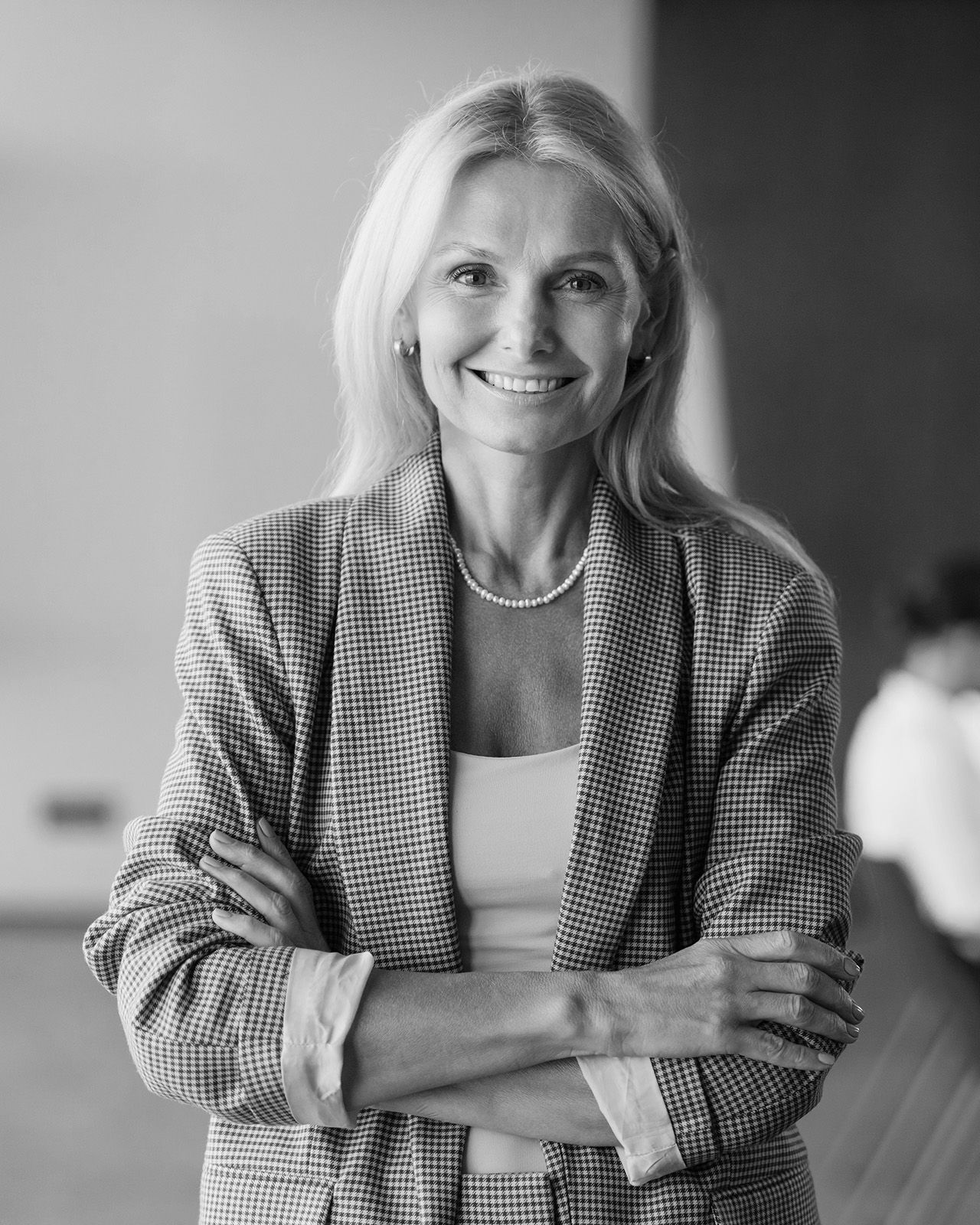 Woman with blonde hair smiles, arms crossed, wearing a blazer, top, and pearl necklace.