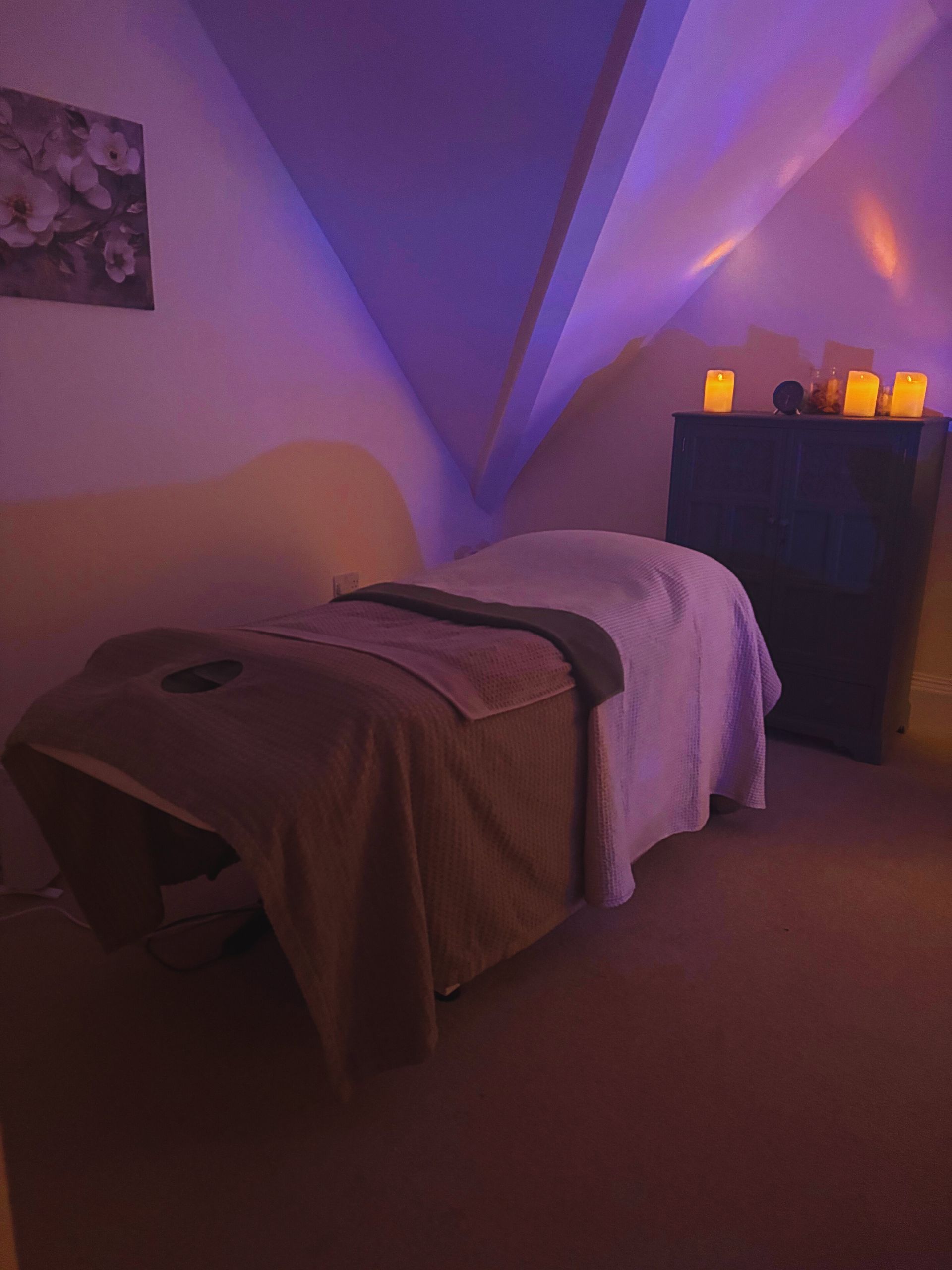 Massage table in a dimly lit room with candles on a dresser under a slanted ceiling, soft purple lighting.