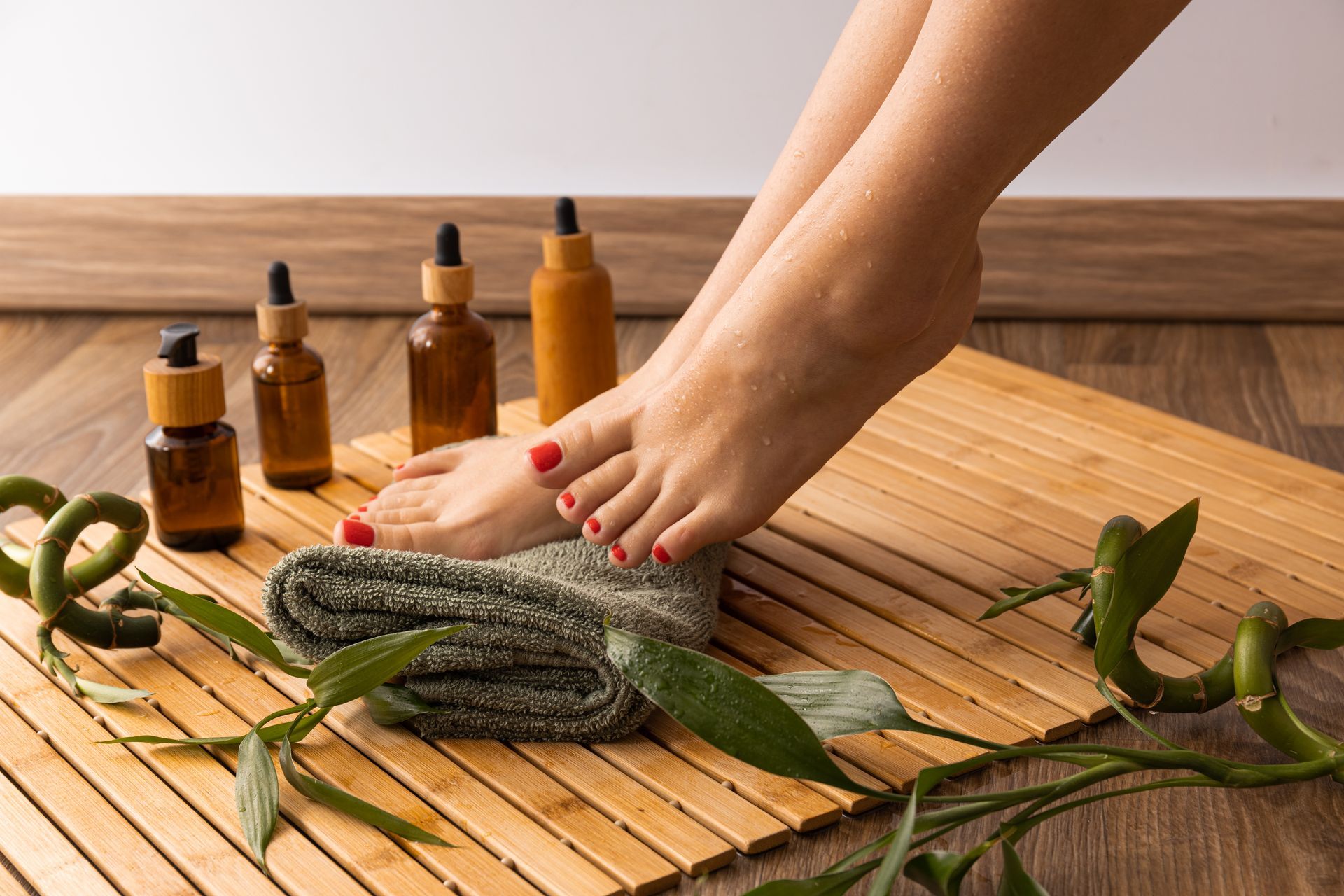 Feet with red nail polish, essential oil bottles, towel, bamboo mat, and plants.