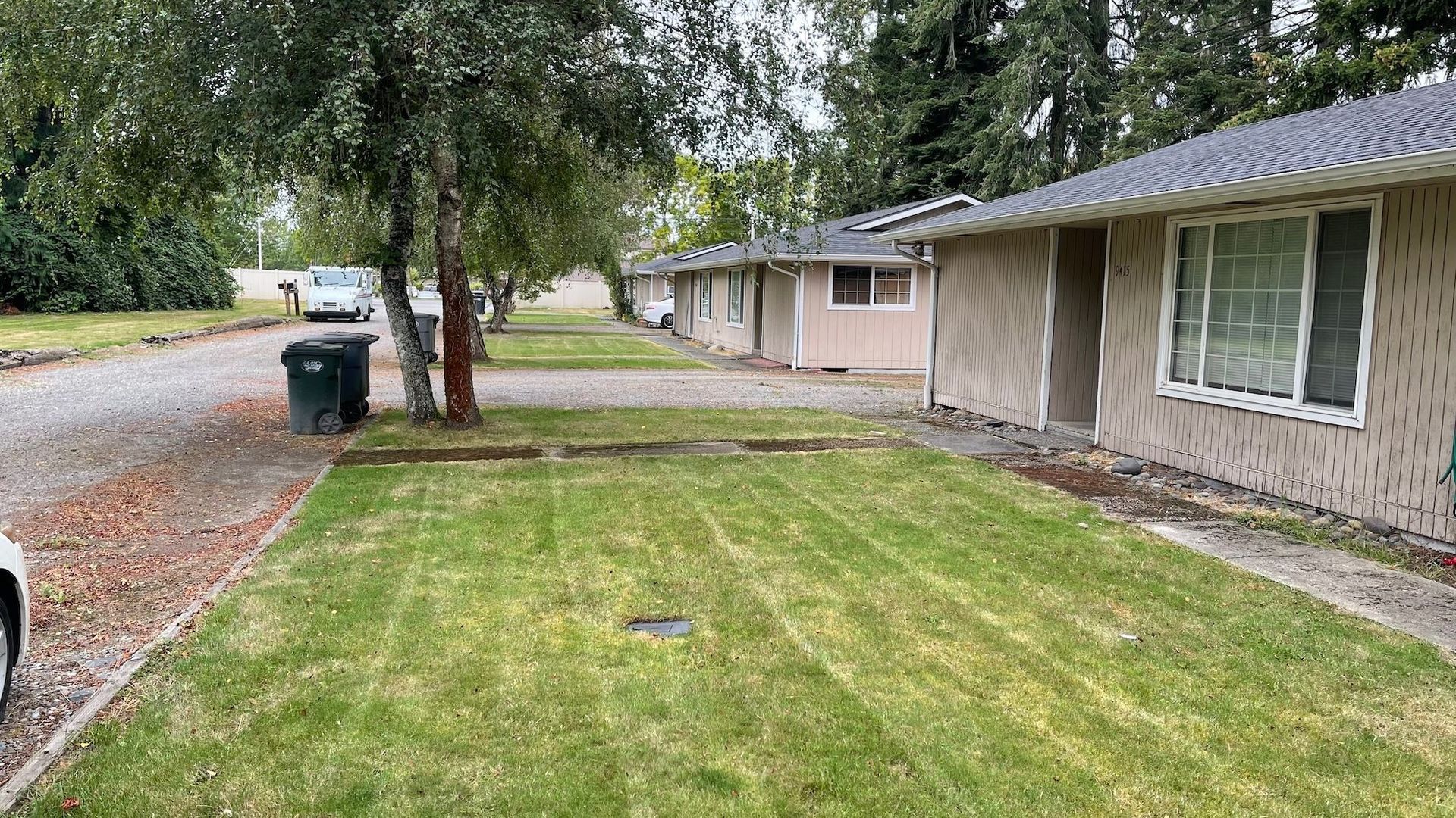 Row of tan, one-story buildings with green lawns and a gravel driveway. Two trash cans and a parked car are visible.