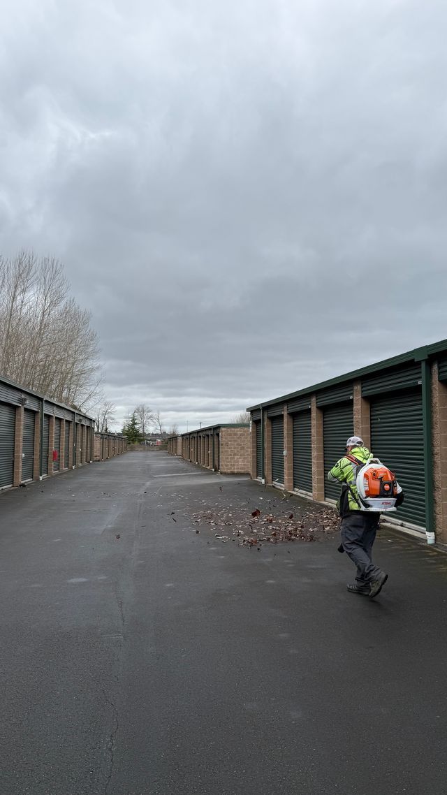 Person using a leaf blower, clearing debris from a storage unit aisle. Gray sky.