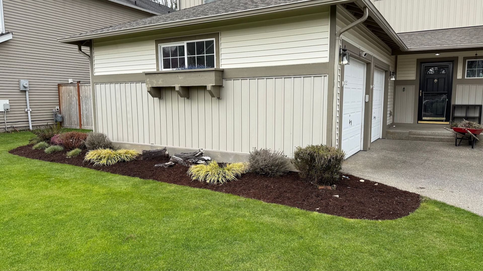 A house with a landscaped garden bed filled with plants and dark mulch, adjacent to a green lawn.