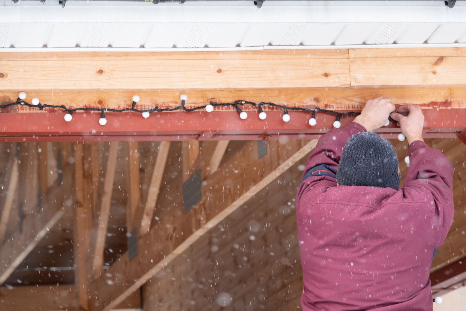Person hangs white globe lights on building's wooden frame in the snow. Person hangs white globe lights on building's wooden frame in the snow.