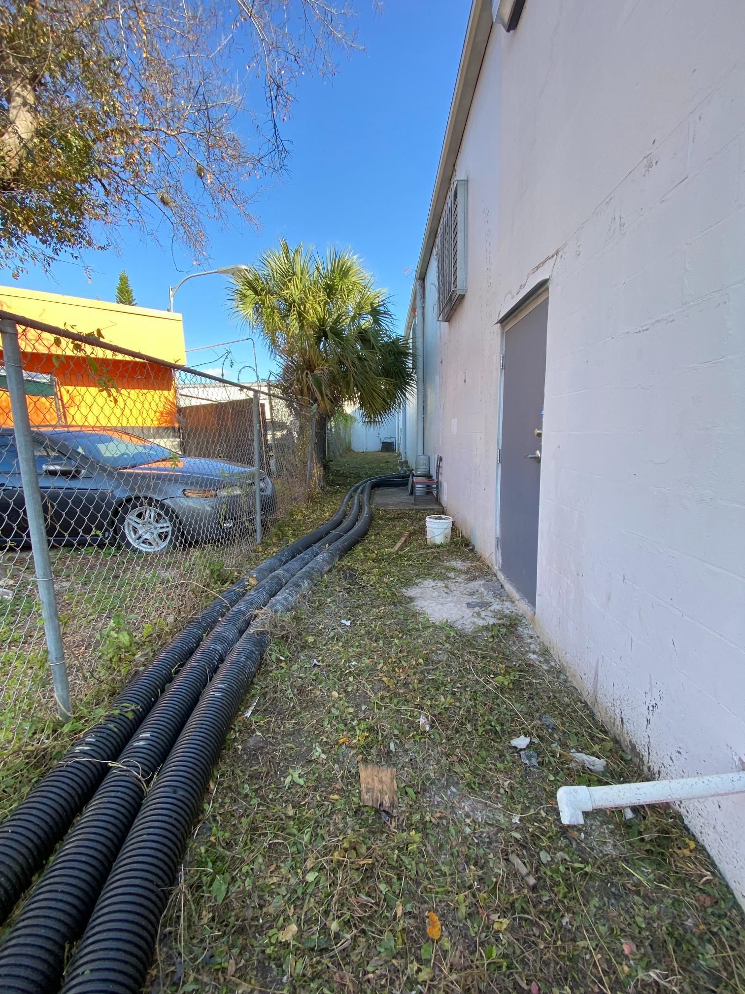Black corrugated pipes run beside a white building and a chain-link fence on a sunny day.