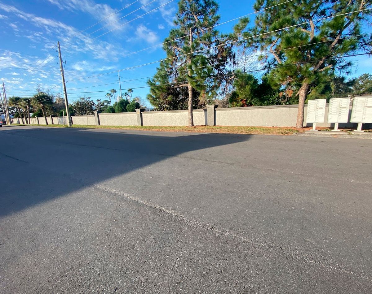 Asphalt road with gray wall and tall trees under a partly cloudy blue sky.