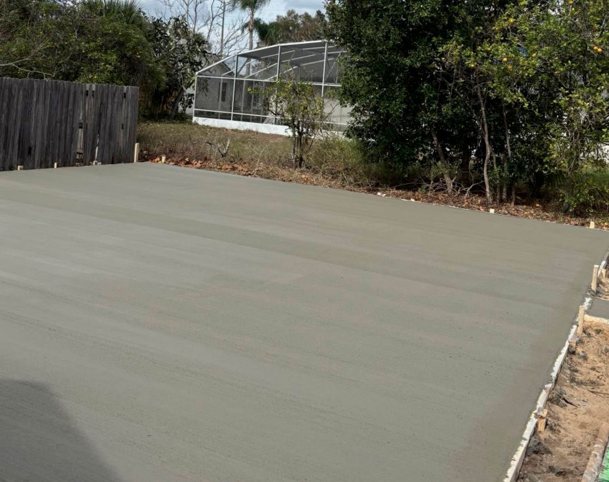 Freshly poured gray concrete driveway. Wooden fence and trees in background.