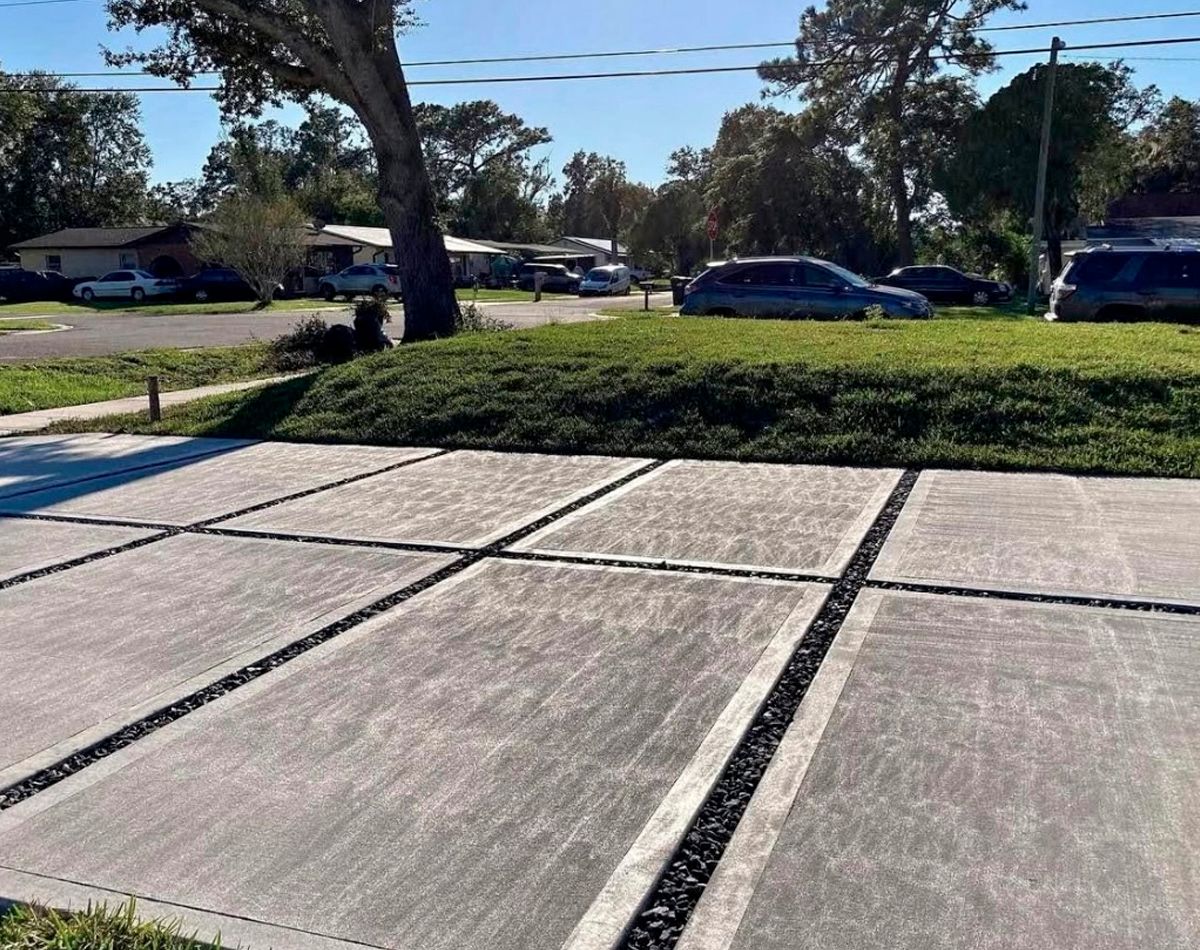 Concrete driveway with dark gravel borders; grassy hill and cars in the background on a sunny day.