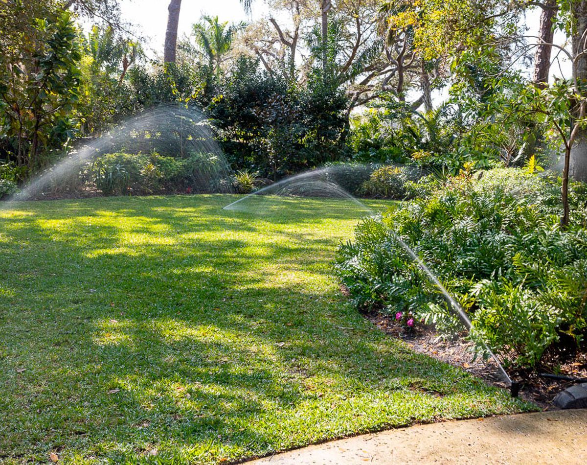 Sprinklers watering a green lawn in a lush garden setting with trees and bushes.
