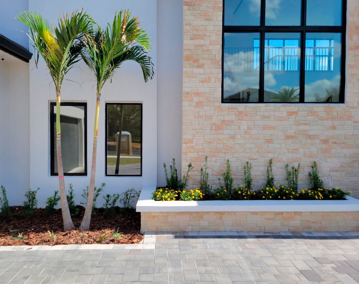 Palm trees next to a modern building with a brick facade and windows, with landscaping and a paver patio.