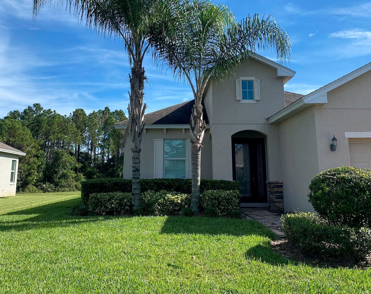 Beige house with palm trees, shrubs, and green lawn under a blue sky.